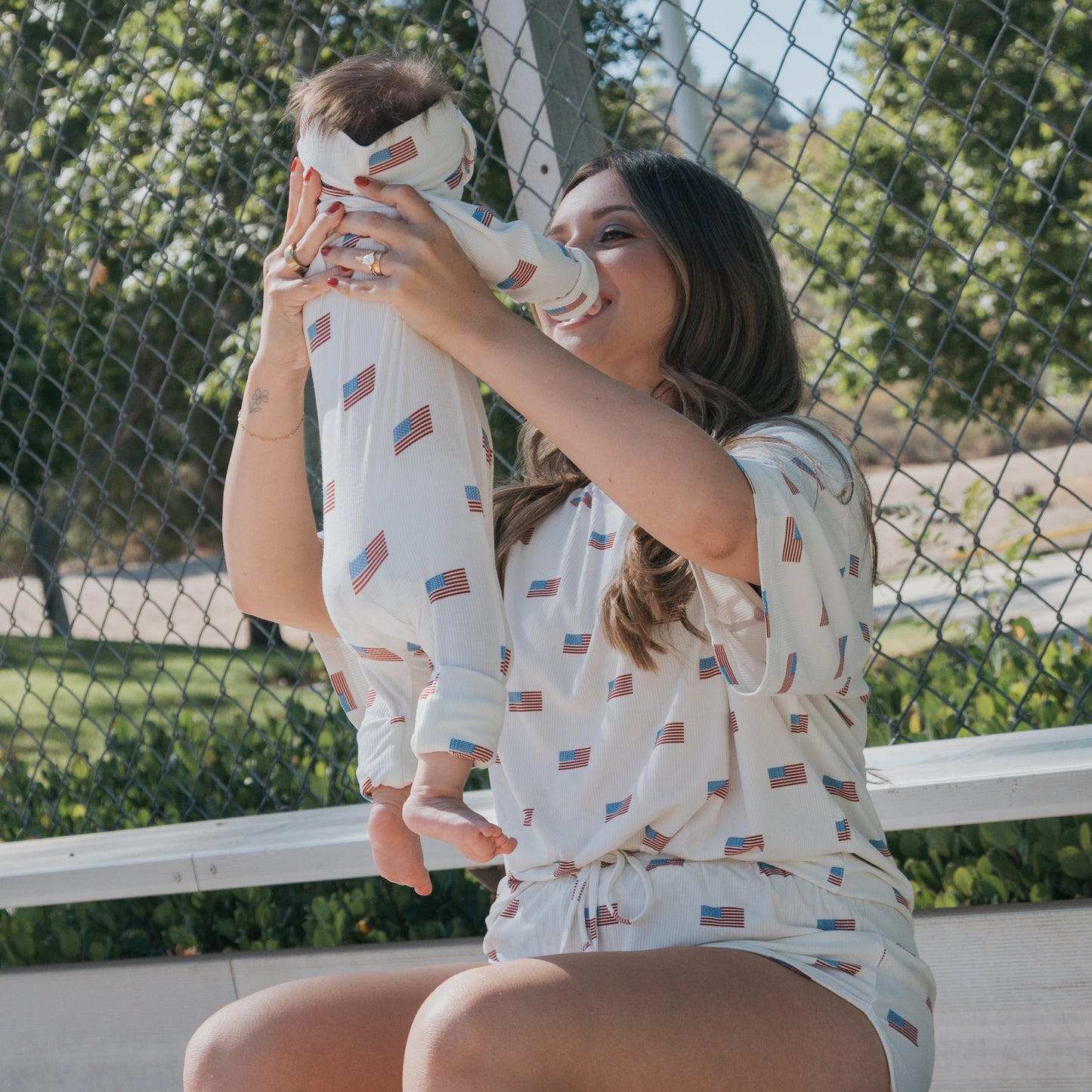 A woman sits on a bench holding a baby in matching forever french baby outfits with American flag prints. The look is completed with the Bamboo Head Wrap | Americana Ribbed by forever french baby. A chain-link fence and trees are in the background.