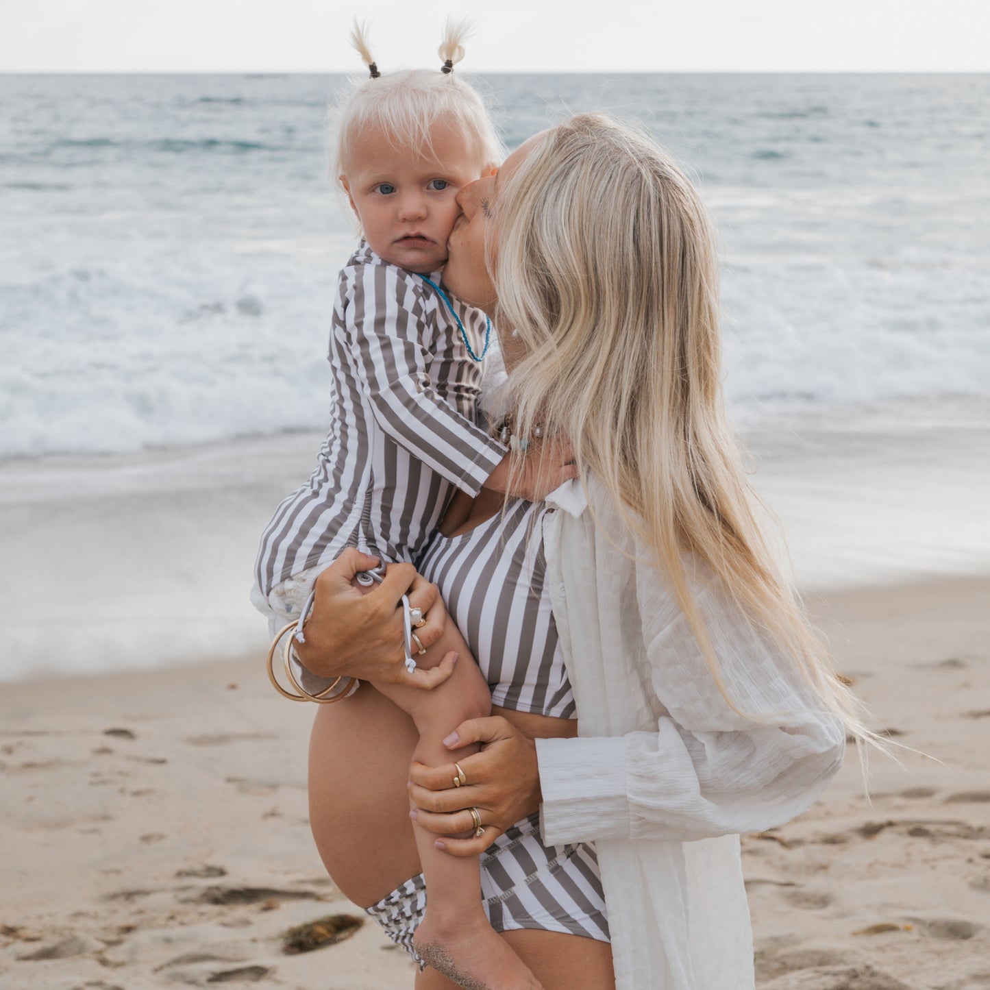 A woman with long blonde hair kisses a young child she holds on a sandy beach. Both wear matching forever french baby Olive Stripe swimsuits, with ocean waves in the background.