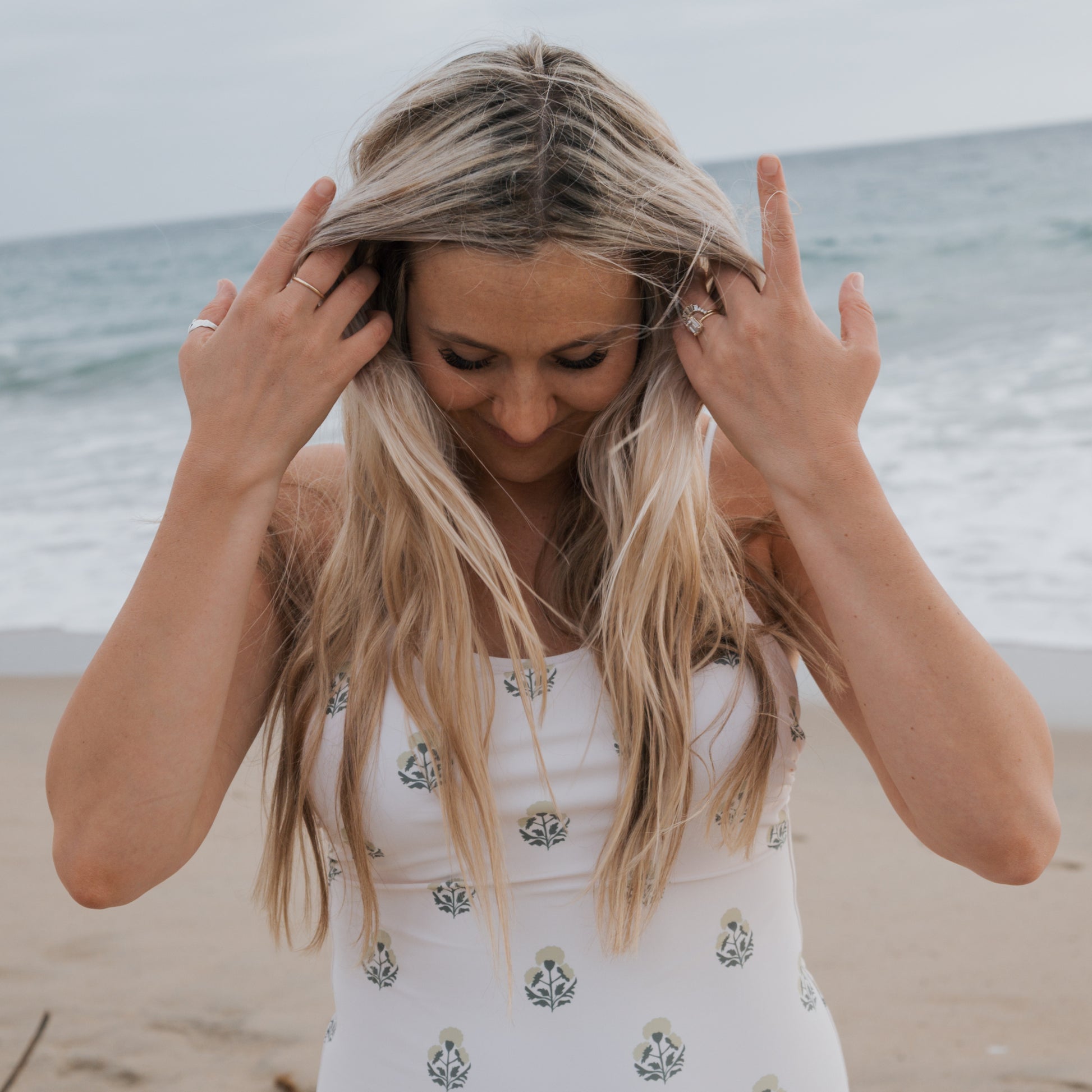 A woman with long blonde hair stands on a sandy beach, adjusting her hair in the Forever French Women's One Piece Swimsuit | Marigold, featuring a white base with green and blue florals, as the ocean provides a serene backdrop.