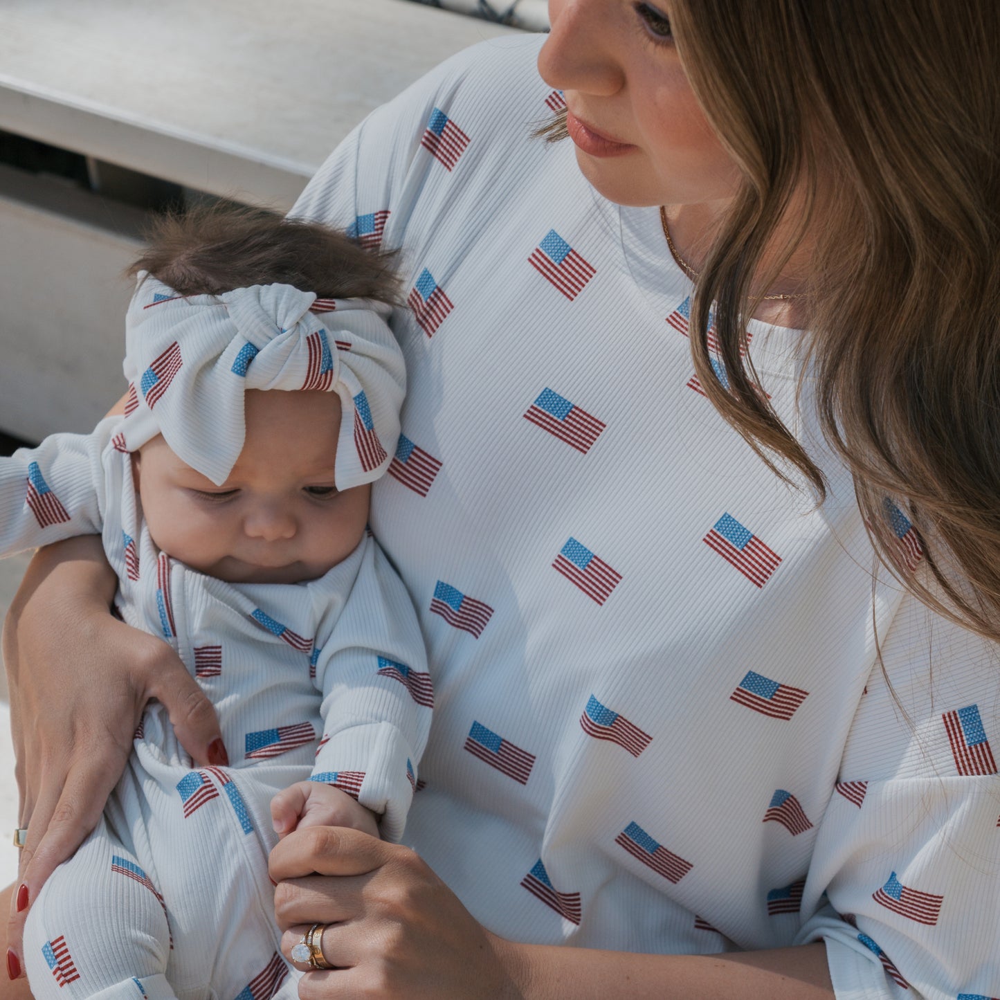A woman gently cradles a baby wearing matching white outfits with American flags. The baby sports the forever french baby Bamboo Head Wrap | Americana Ribbed, adding cozy, stylish flair to their look.