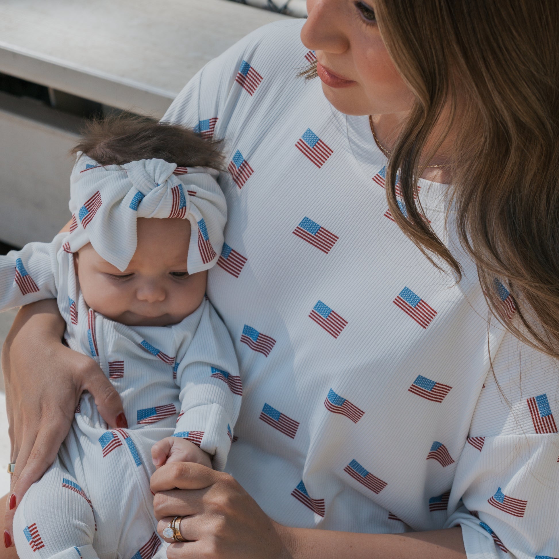 A woman gently cradles a baby wearing matching white outfits with American flags. The baby sports the forever french baby Bamboo Head Wrap | Americana Ribbed, adding cozy, stylish flair to their look.