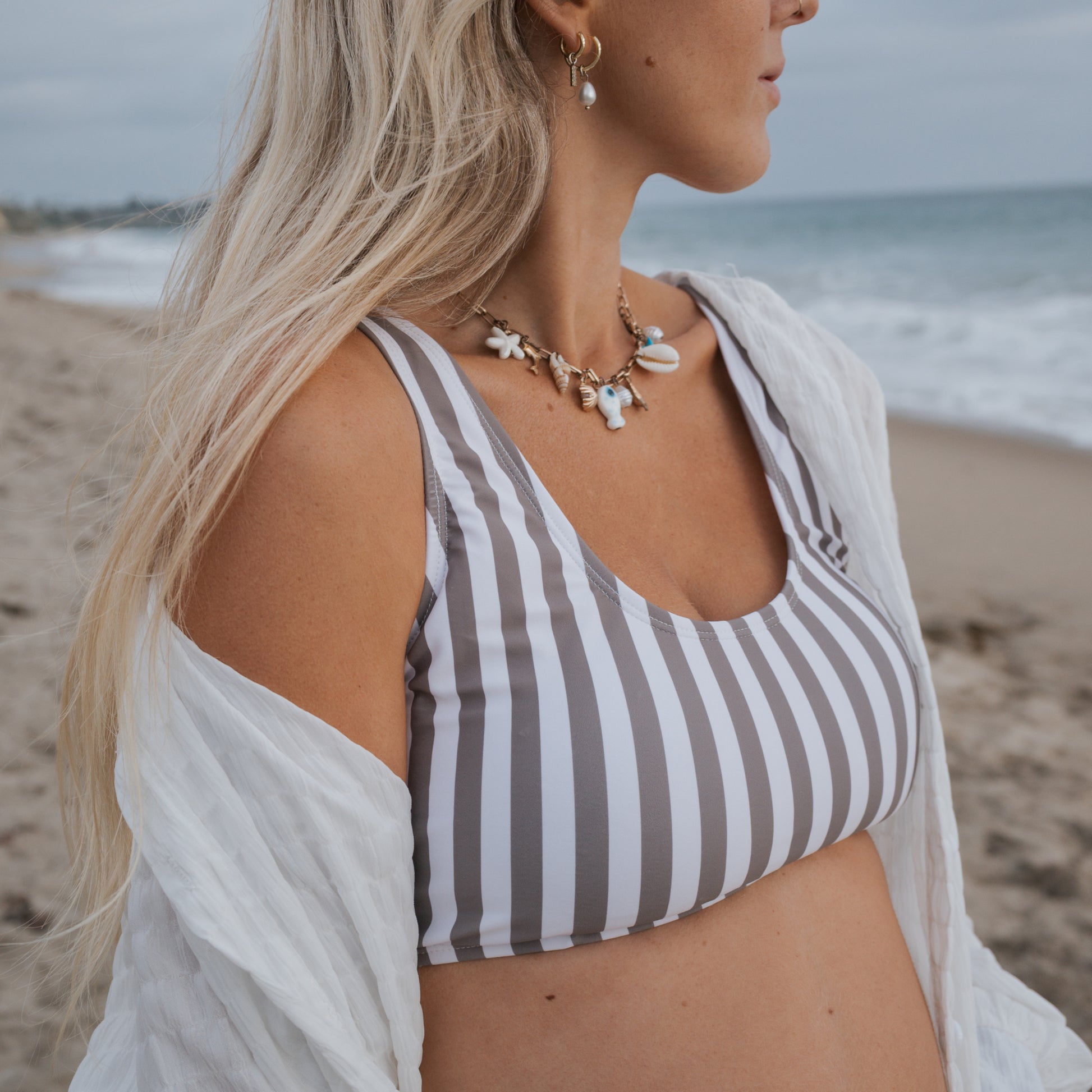 A woman with long blonde hair stands on the beach in the forever french baby Women's Two Piece Swimsuit | Olive Stripe, styled with a white cover-up and seashell necklace. The ocean and cloudy sky are behind her, face partly out of frame.