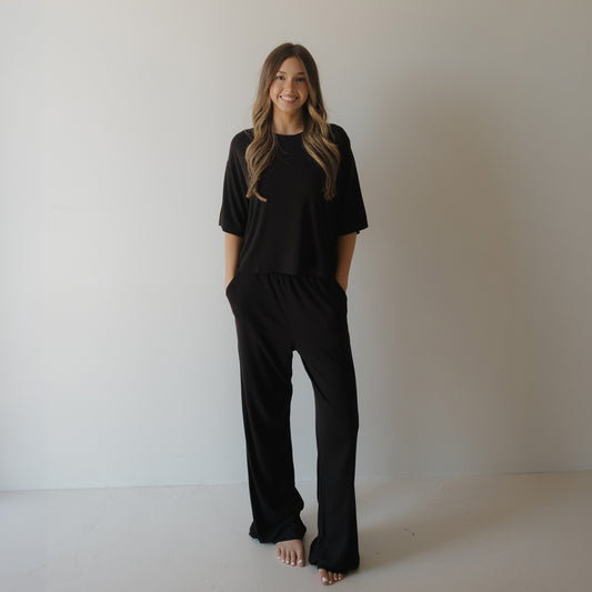 A woman with long wavy hair, barefoot and smiling with hands in her pockets, wears the Women's Boxy Tee Lounge Set | Ribbed Black by forever french baby as she stands against a plain white wall.