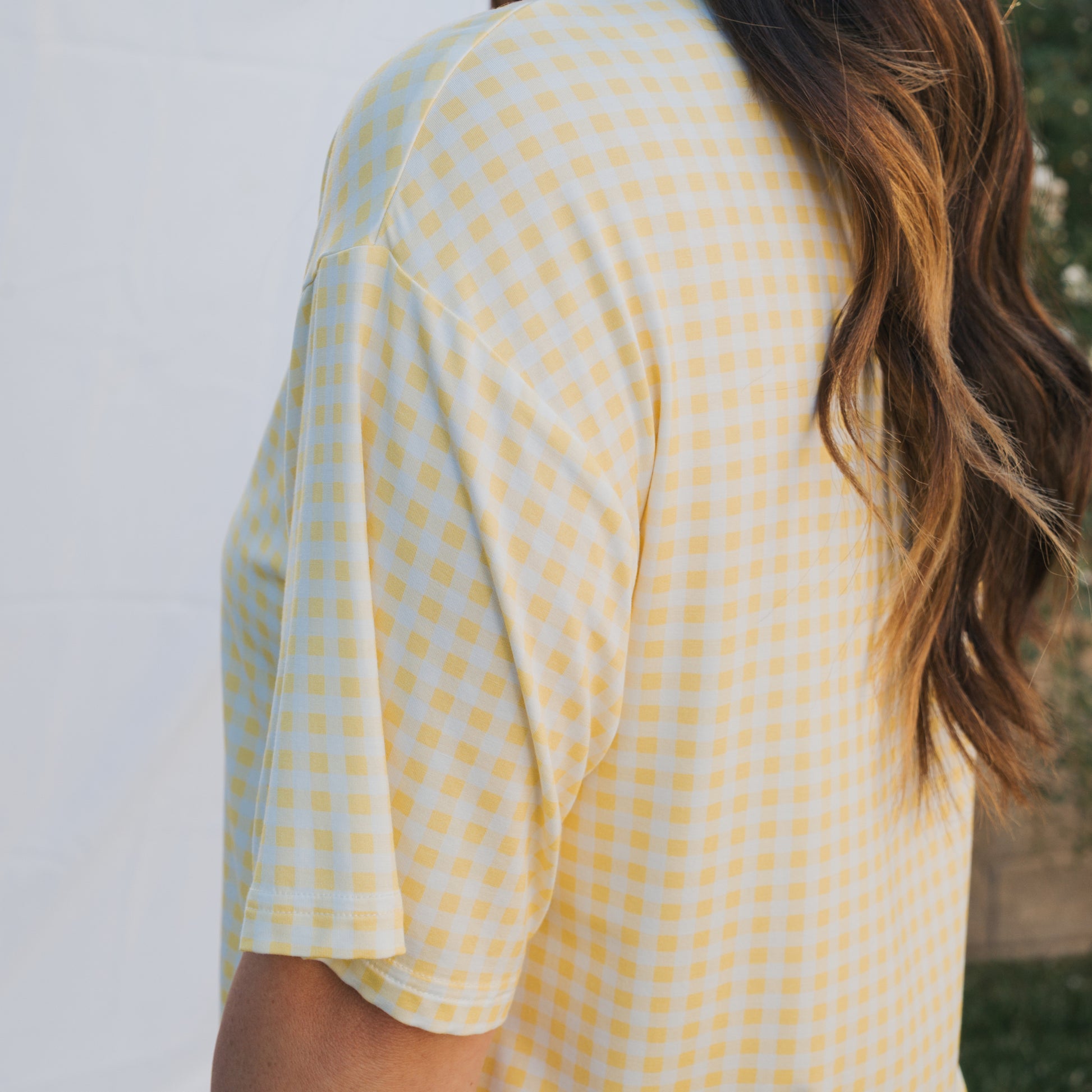 A woman with long, wavy hair wears the forever french baby Women's Bamboo Short Set in Citron, a yellow and white checkered, breathable outfit. She is shown from the back and side outdoors among white fabric and greenery.