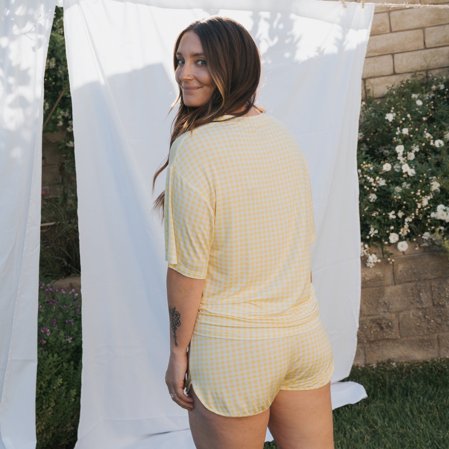 A woman with long brown hair stands outdoors, smiling over her shoulder in the forever french baby Women's Bamboo Short Set | Citron. She is surrounded by greenery, a white sheet, and a brick wall in the background.