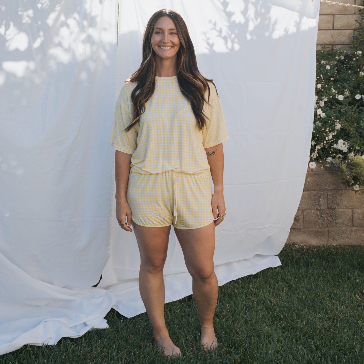 A woman stands barefoot on grass before a white sheet, smiling with long wavy hair. She wears the forever french baby Women's Bamboo Short Set in Citron—a breathable, stylish, and comfortable summer essential.