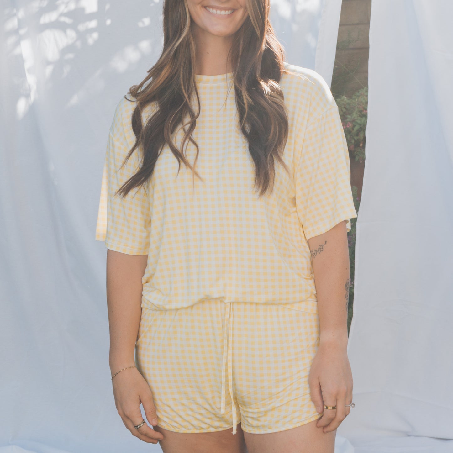 A woman stands outdoors, smiling with her hair down, wearing the forever french baby Women's Bamboo Short Set in Citron—featuring a yellow and white gingham short-sleeve top and matching shorts—against a backdrop of greenery.