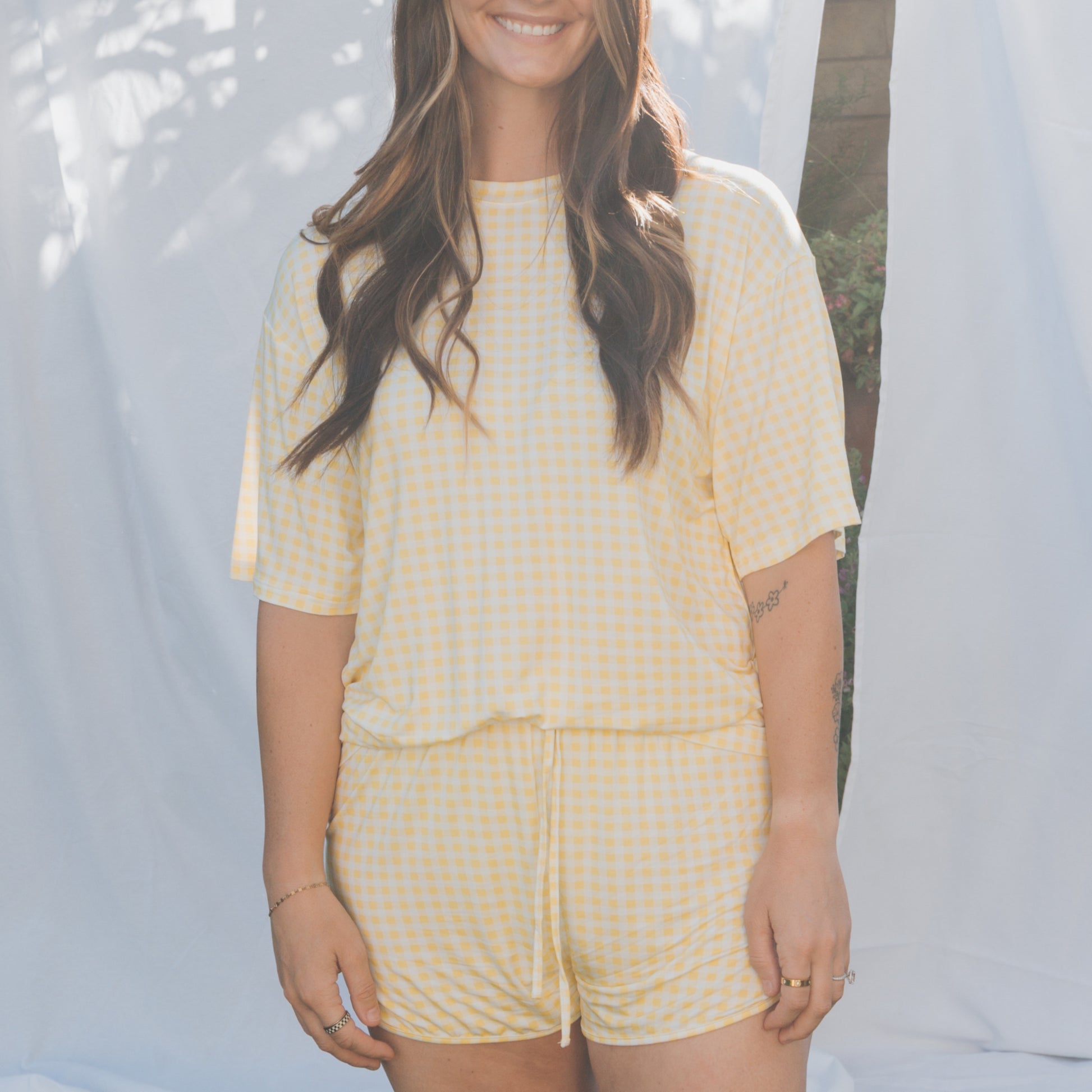 A woman stands outdoors, smiling with her hair down, wearing the forever french baby Women's Bamboo Short Set in Citron—featuring a yellow and white gingham short-sleeve top and matching shorts—against a backdrop of greenery.
