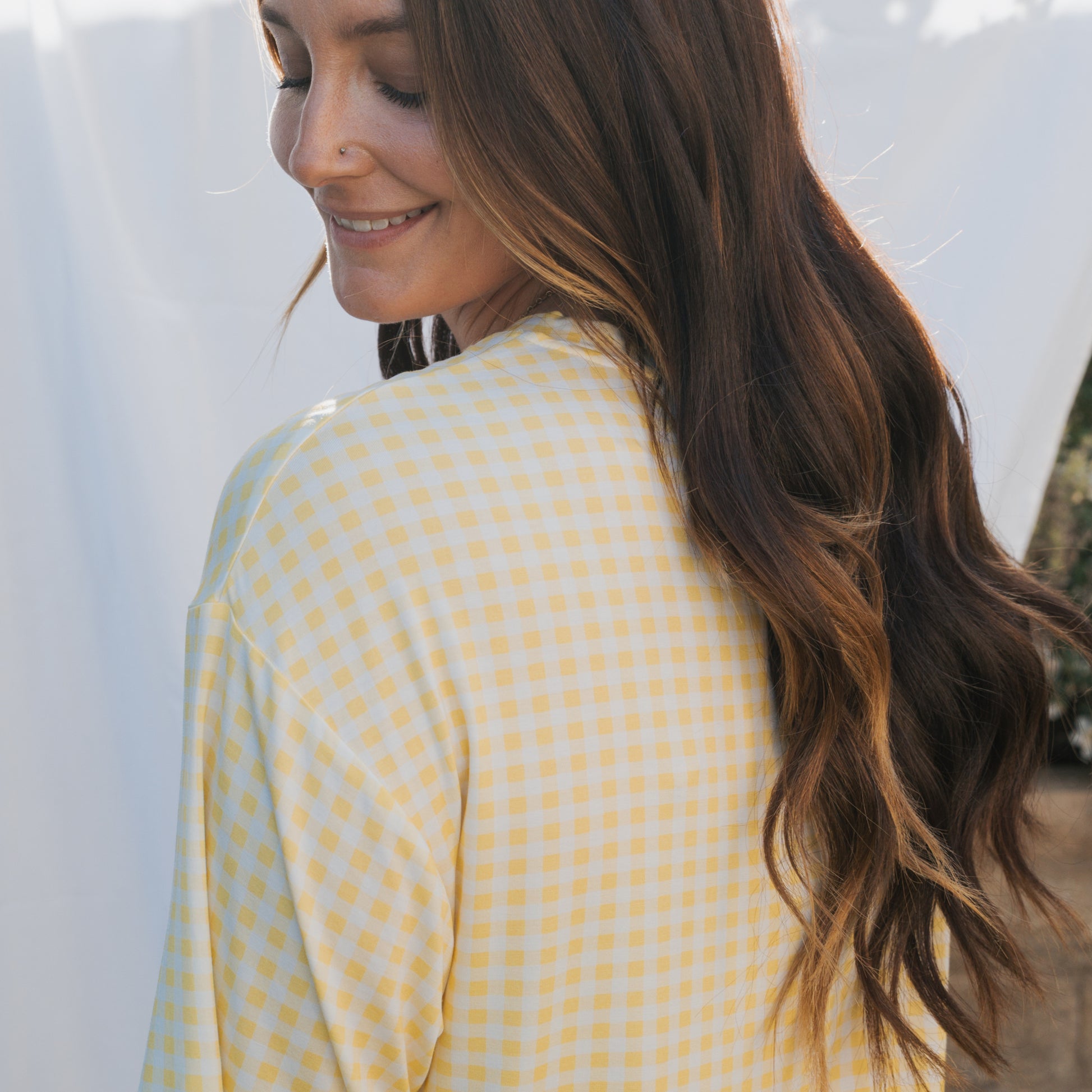 A woman with long, wavy brown hair smiles over her shoulder, wearing the forever french baby Women's Bamboo Short Set in Citron against a softly lit background.