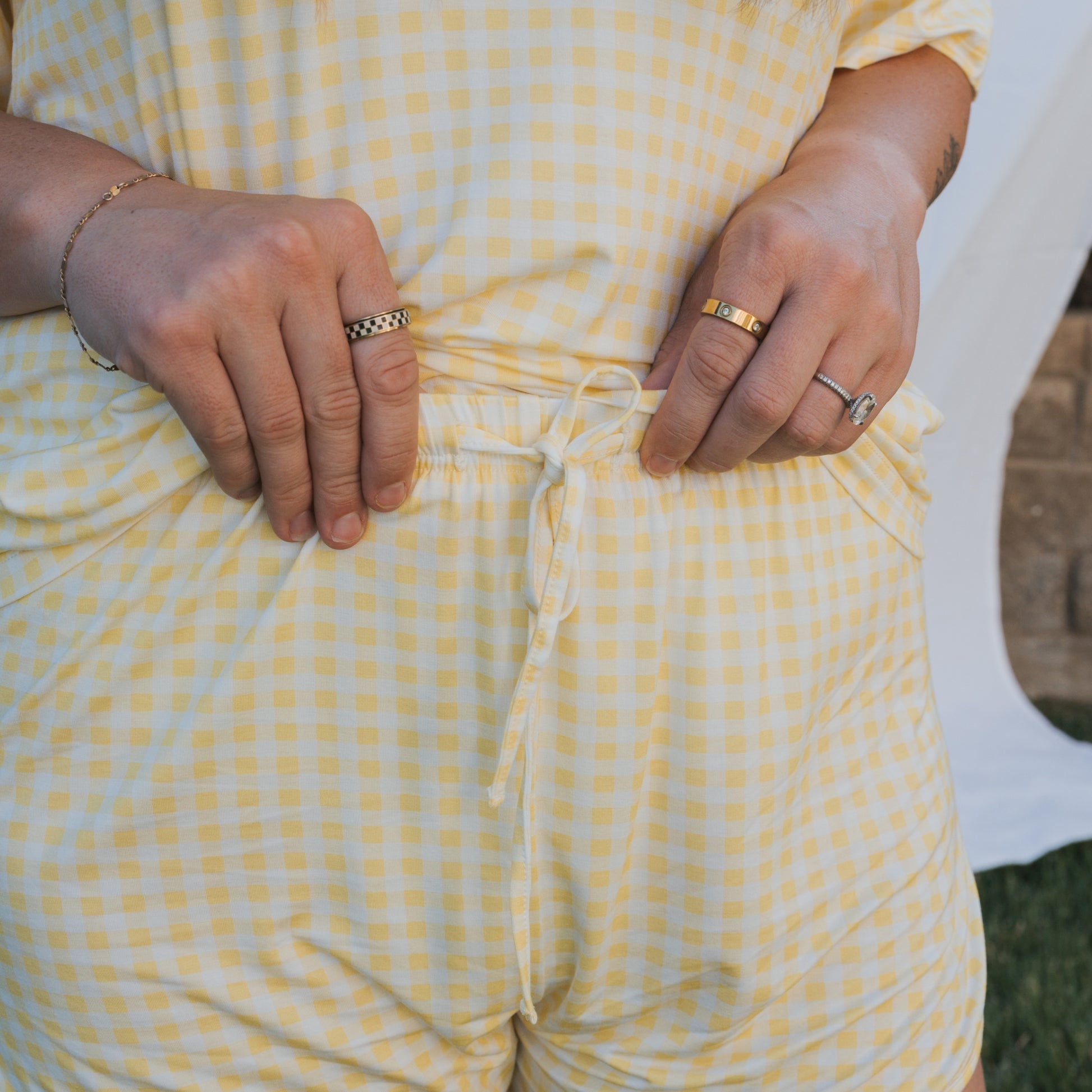 Close-up of a person adjusting the drawstring on forever french baby's Women's Bamboo Short Set | Citron, featuring yellow and white gingham shorts and a matching top, accessorized with rings and bracelets. Outdoor backdrop partially visible.