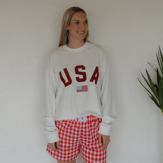 A woman smiles and looks to the side, standing by a green plant in a relaxed white "USA" sweatshirt paired with forever french baby's Women's Boxer Shorts | Scarlet Gingham against a plain white wall.
