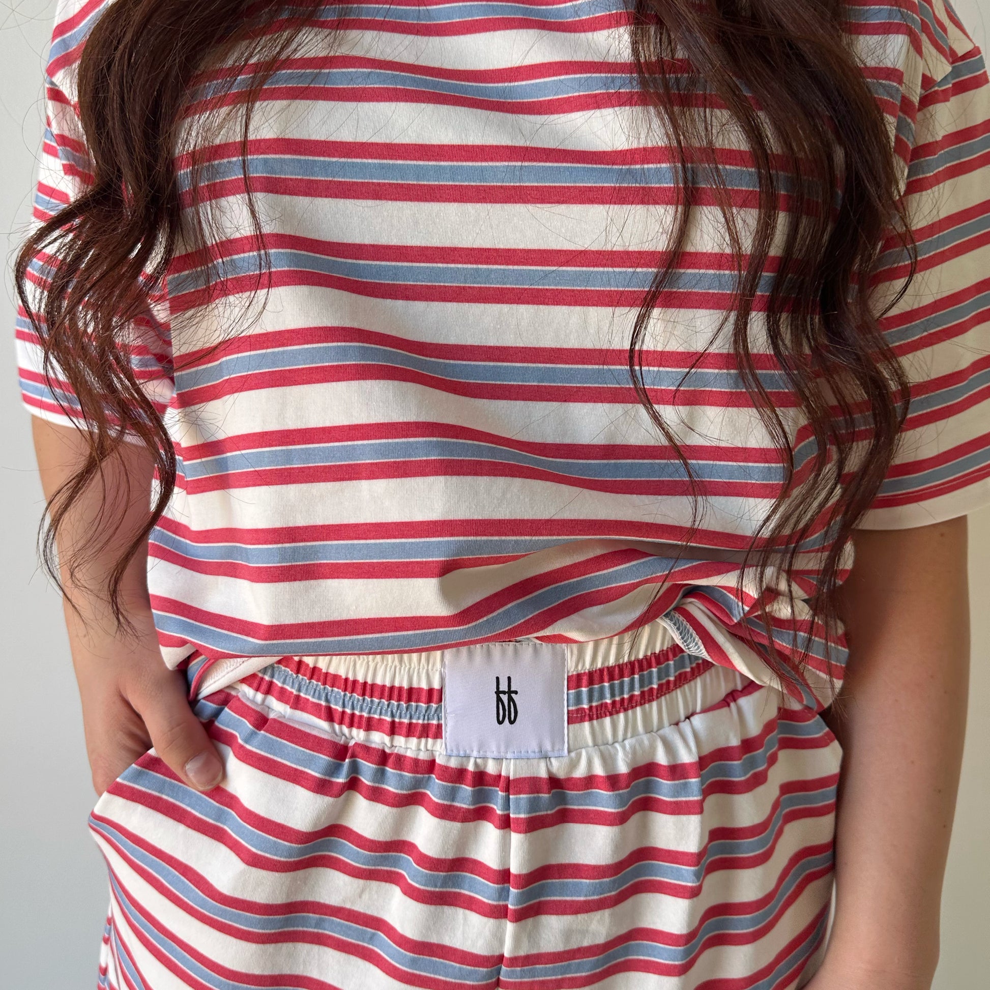 A person wears the forever french baby Women's Short Set | Heritage Stripe in red, blue, and white. Their long wavy brown hair flows over the matching shirt and shorts, which have a white label with a small black logo on the waistband.