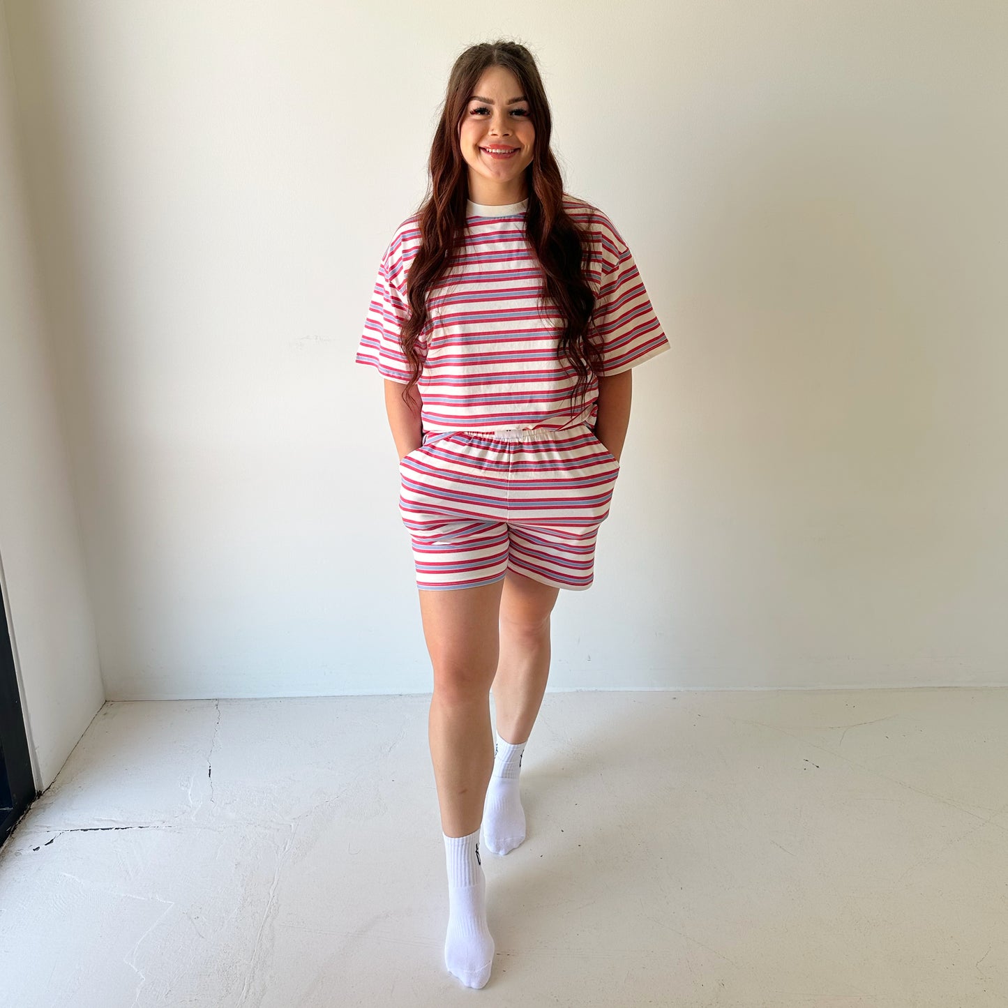 A woman smiles indoors in the forever french baby Women's Short Set | Heritage Stripe, featuring relaxed-fit pink and white stripes, hands in her pockets, and white socks against a minimal plain background.