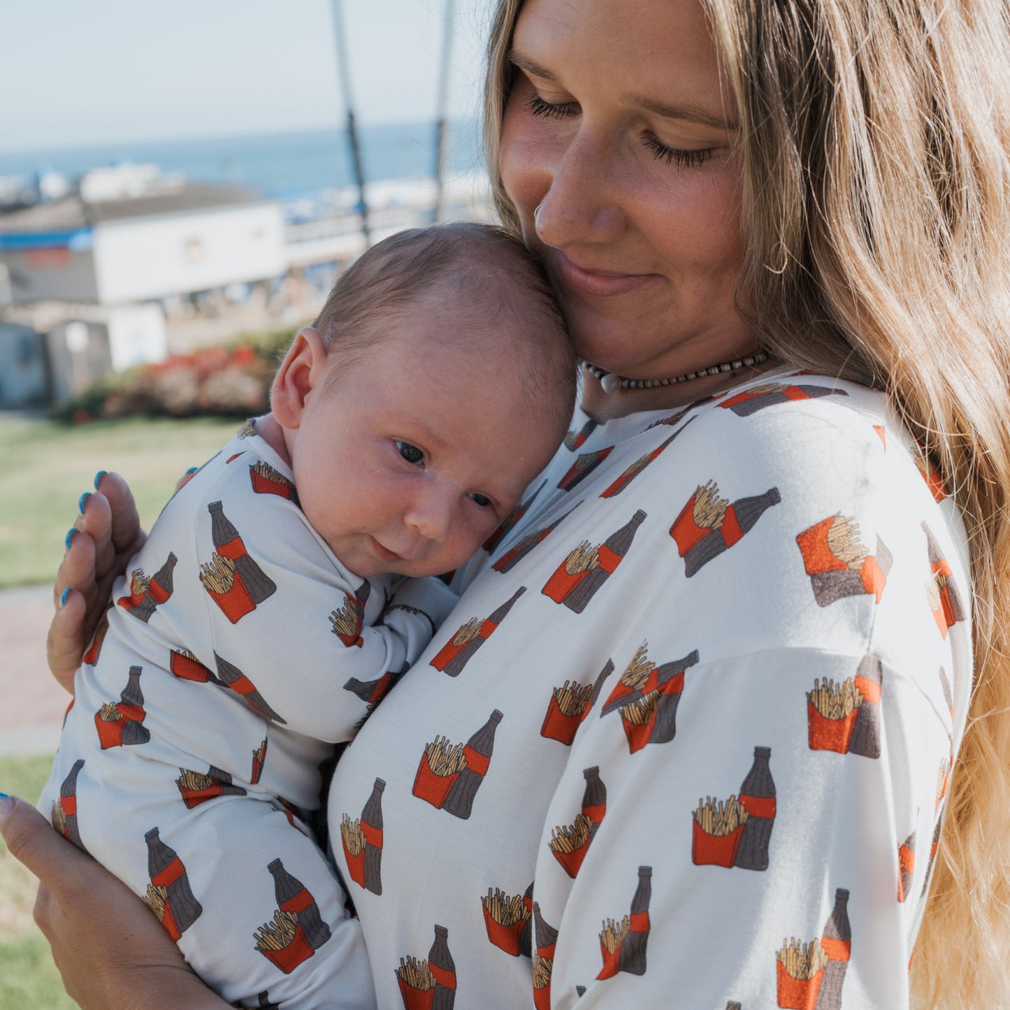 A woman and baby wear matching forever french baby Women's Bamboo Short Sets in the Fries & Cola print, showcasing stylish, hypo-allergenic outfits with a fun soda and fries pattern. They're outside on a sunny day with blurred buildings and ocean behind.