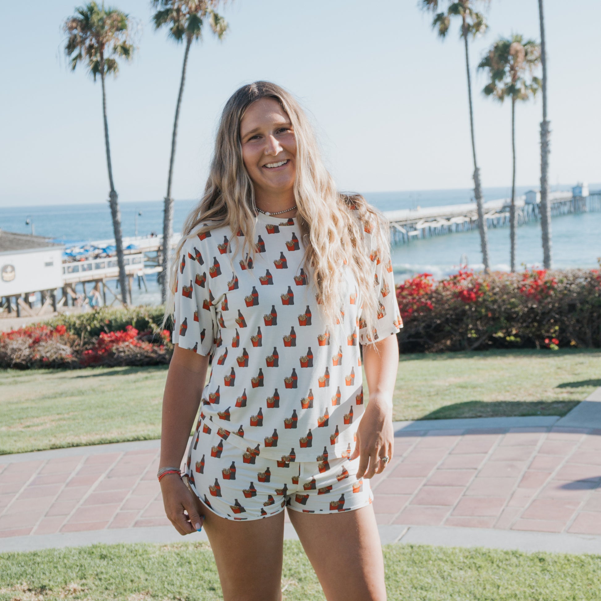 A woman with long, wavy hair smiles outdoors by the ocean, wearing forever french baby's Women's Bamboo Short Set in the Fries & Cola print. Palm trees and a pier are visible in the sunny background.