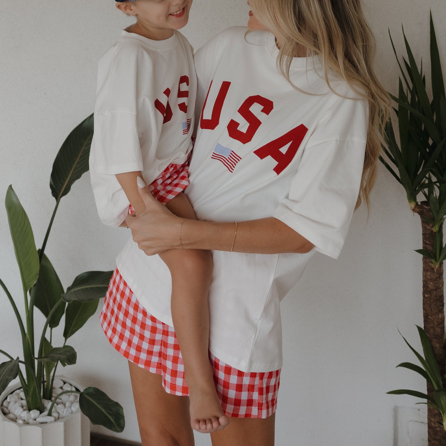 A woman and a young child stand indoors by green potted plants, both wearing matching white USA shirts and forever french baby's Women's Boxer Shorts | Scarlet Gingham, made from 100% cotton.