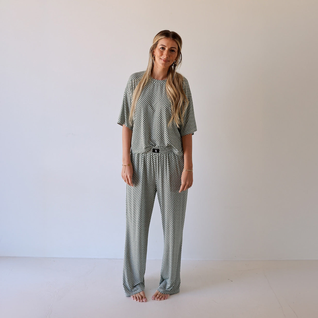 A woman with long, wavy hair stands barefoot against a light wall, smiling gently and wearing the forever french baby Women's Boxy Tee Lounge Set in Lucky Micro Check. Her arms are relaxed as she models the loose-fitting outfit.