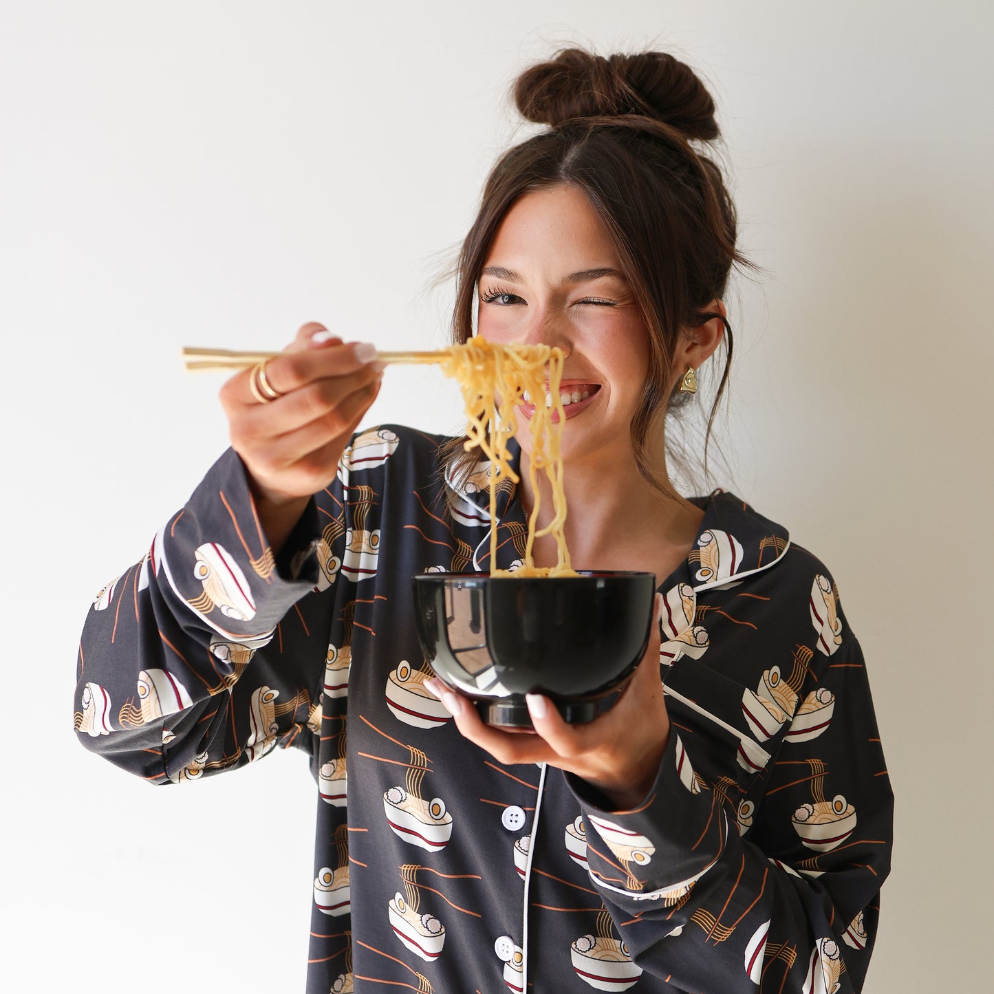 A smiling woman wearing the Women's Bamboo Button Up Lounge Set | Ramen by forever french baby holds a black bowl of noodles and eats with chopsticks, winking playfully at the camera against a plain white background.