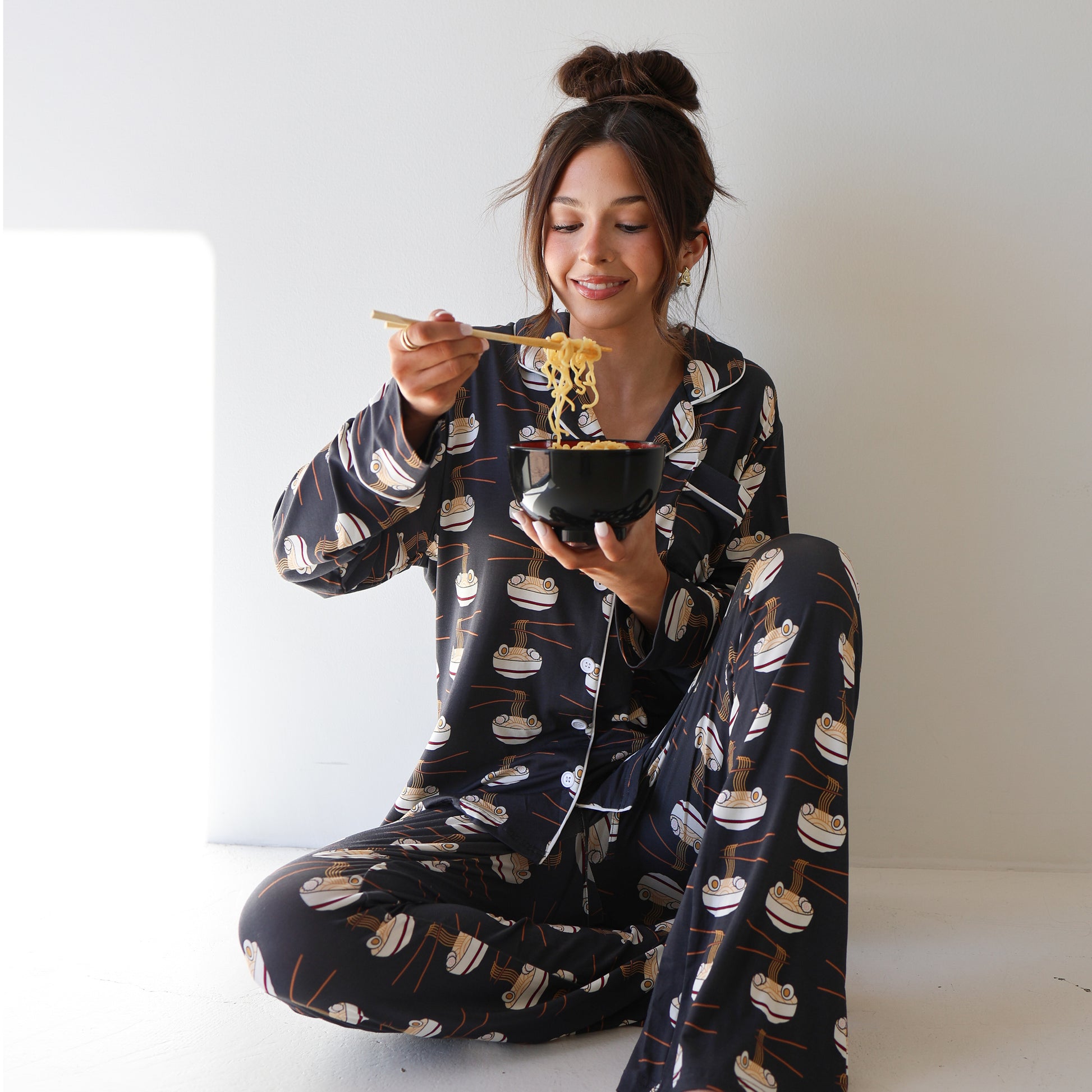 A woman in the forever french baby Women's Bamboo Button Up Lounge Set | Ramen sits on the floor, smiling as she lifts noodles with chopsticks, enjoying the soft, hypo-allergenic comfort of her bamboo loungewear.
