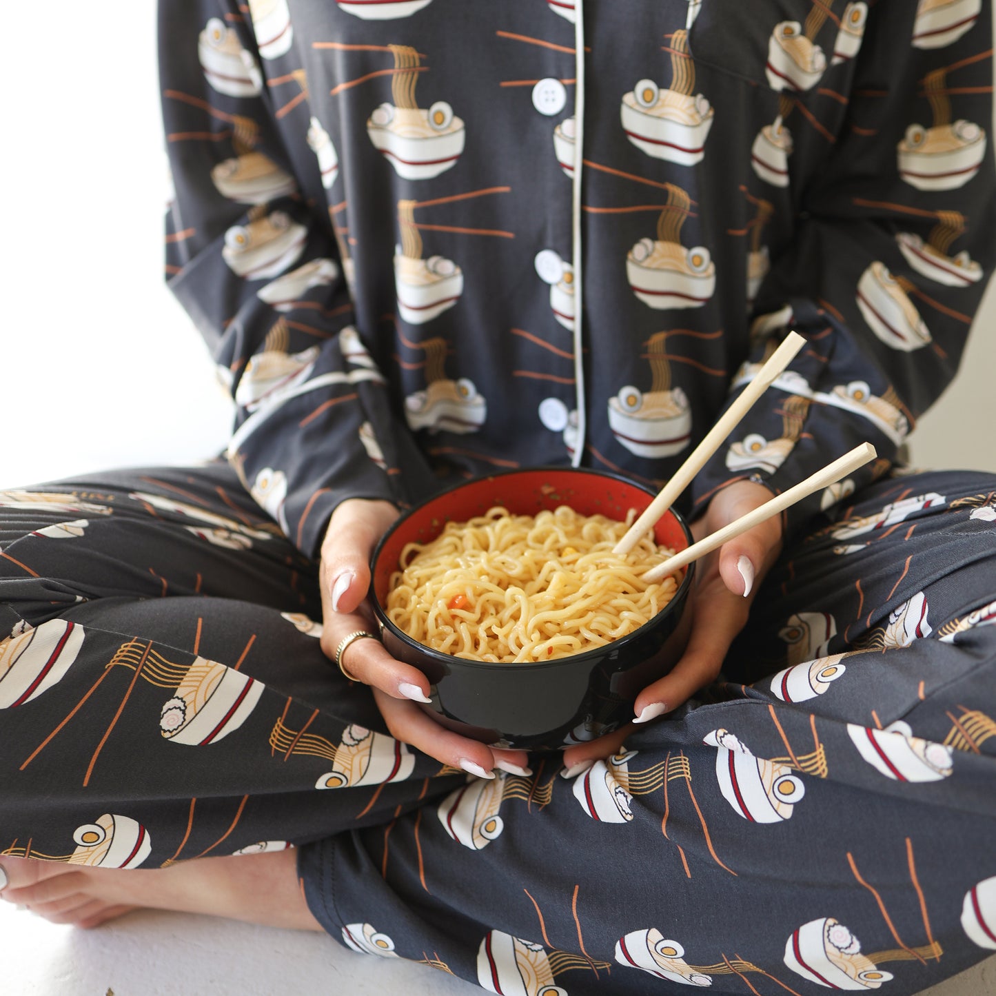 A person in the forever french baby Women's Bamboo Button Up Lounge Set | Ramen sits cross-legged, enjoying ramen with chopsticks. The hypoallergenic bamboo fabric enhances their comfort while they relax.