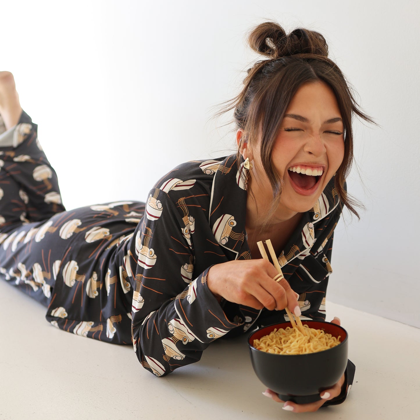 A woman in the forever french baby Women's Bamboo Button Up Lounge Set | Ramen lies on the floor, eyes closed and laughing, as she uses chopsticks to eat noodles from a black and red bowl.