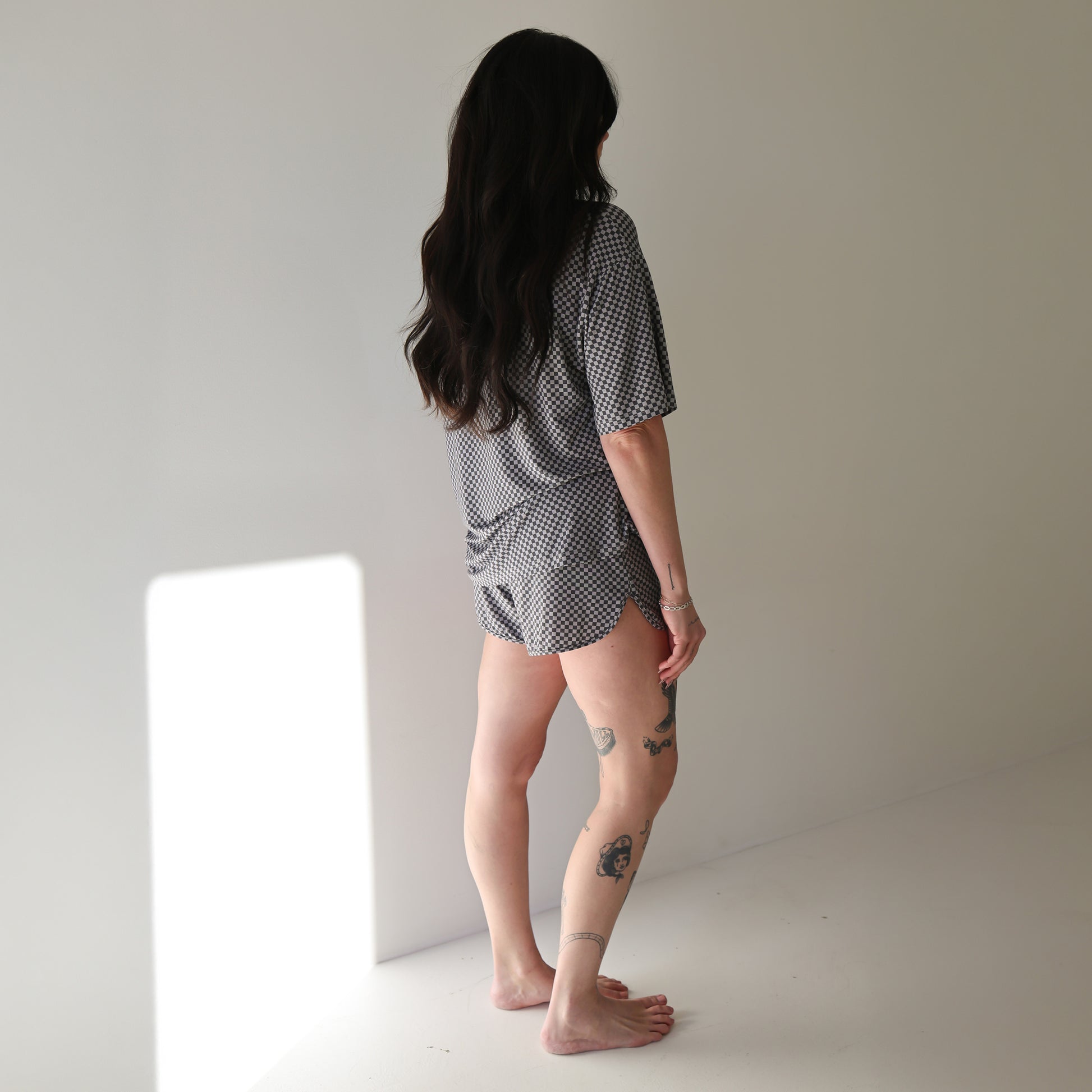 A person with long dark hair stands barefoot in a sunlit room, facing away from the camera. They wear the forever french baby Women's Bamboo Short Set | Street Micro Check and have several tattoos on their arms and legs.
