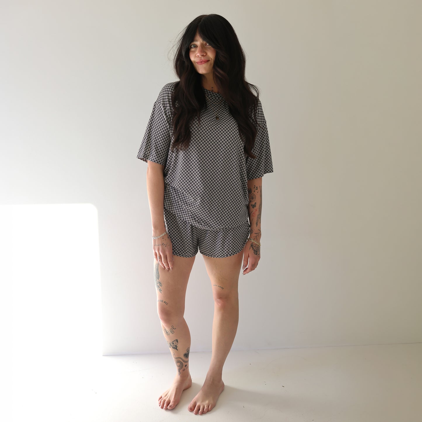 A woman with long dark hair and visible tattoos stands barefoot in a well-lit room, smiling slightly while wearing the forever french baby Women's Bamboo Short Set | Street Micro Check.