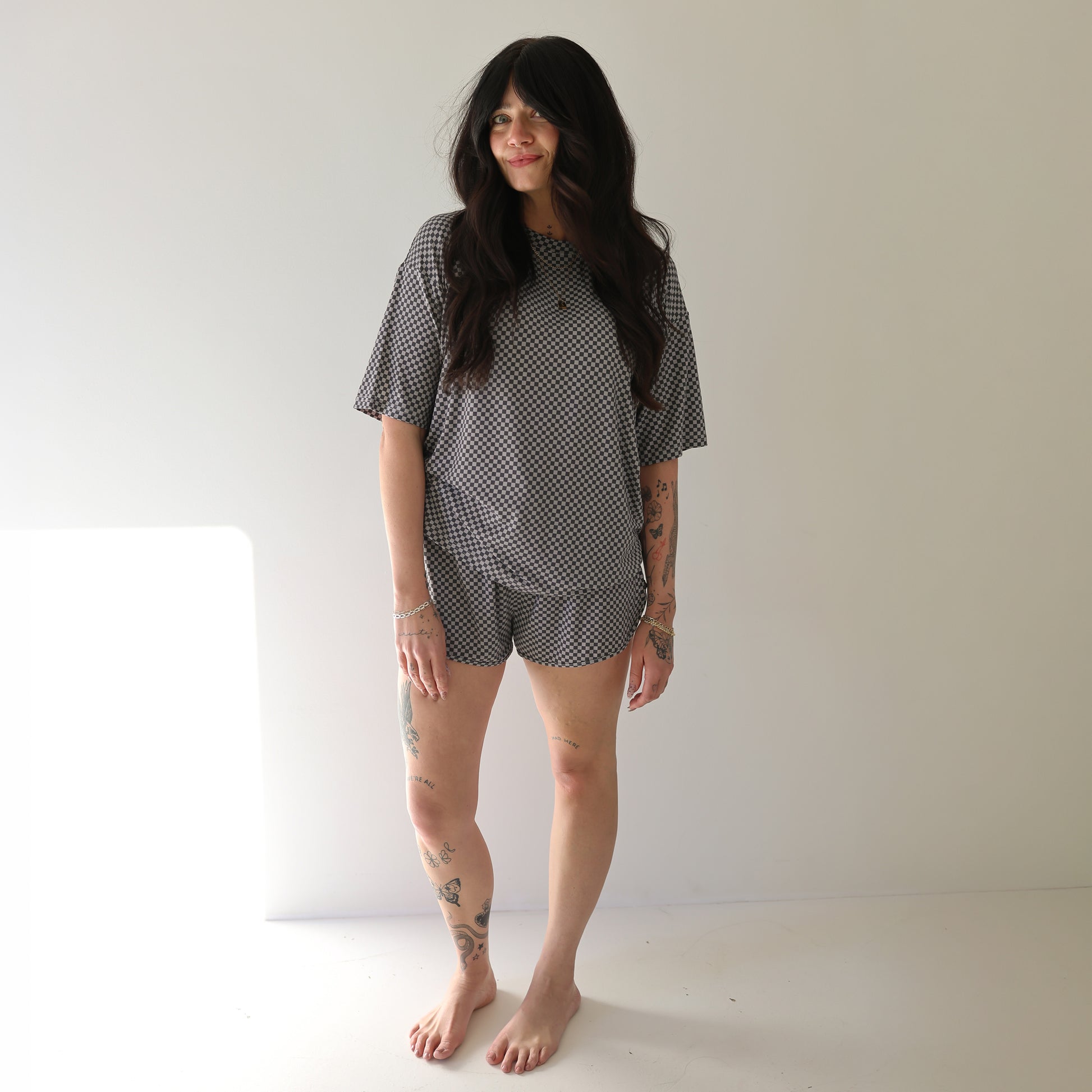 A woman with long dark hair and visible tattoos stands barefoot in a well-lit room, smiling slightly while wearing the forever french baby Women's Bamboo Short Set | Street Micro Check.