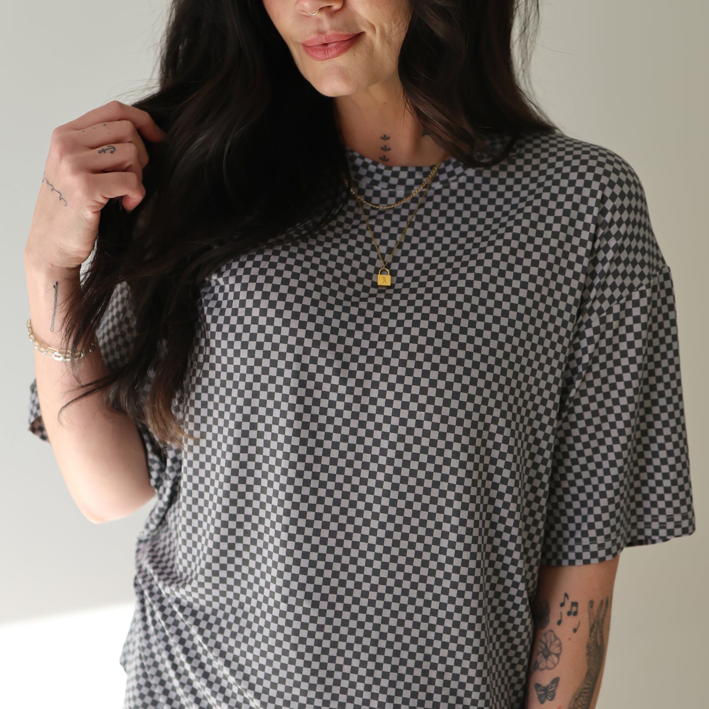 A woman with long dark hair and tattoos wears the forever french baby Women's Bamboo Short Set in Street Micro Check, pairing it with gold necklaces. She stands against a plain light background, holding her hair with one hand.