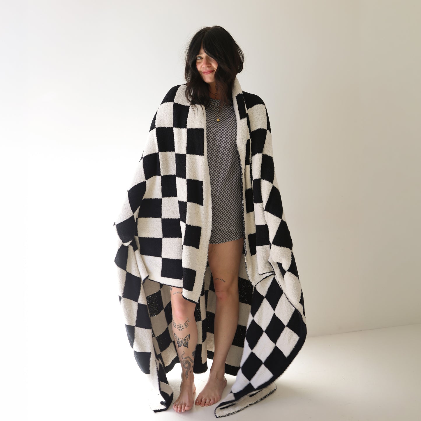 A woman, barefoot in a bright room, is wrapped in the forever french baby Plush Blanket | Black & White Checkerboard. She wears a short patterned dress, has tattoos on her legs, and smiles gently while looking to the side.
