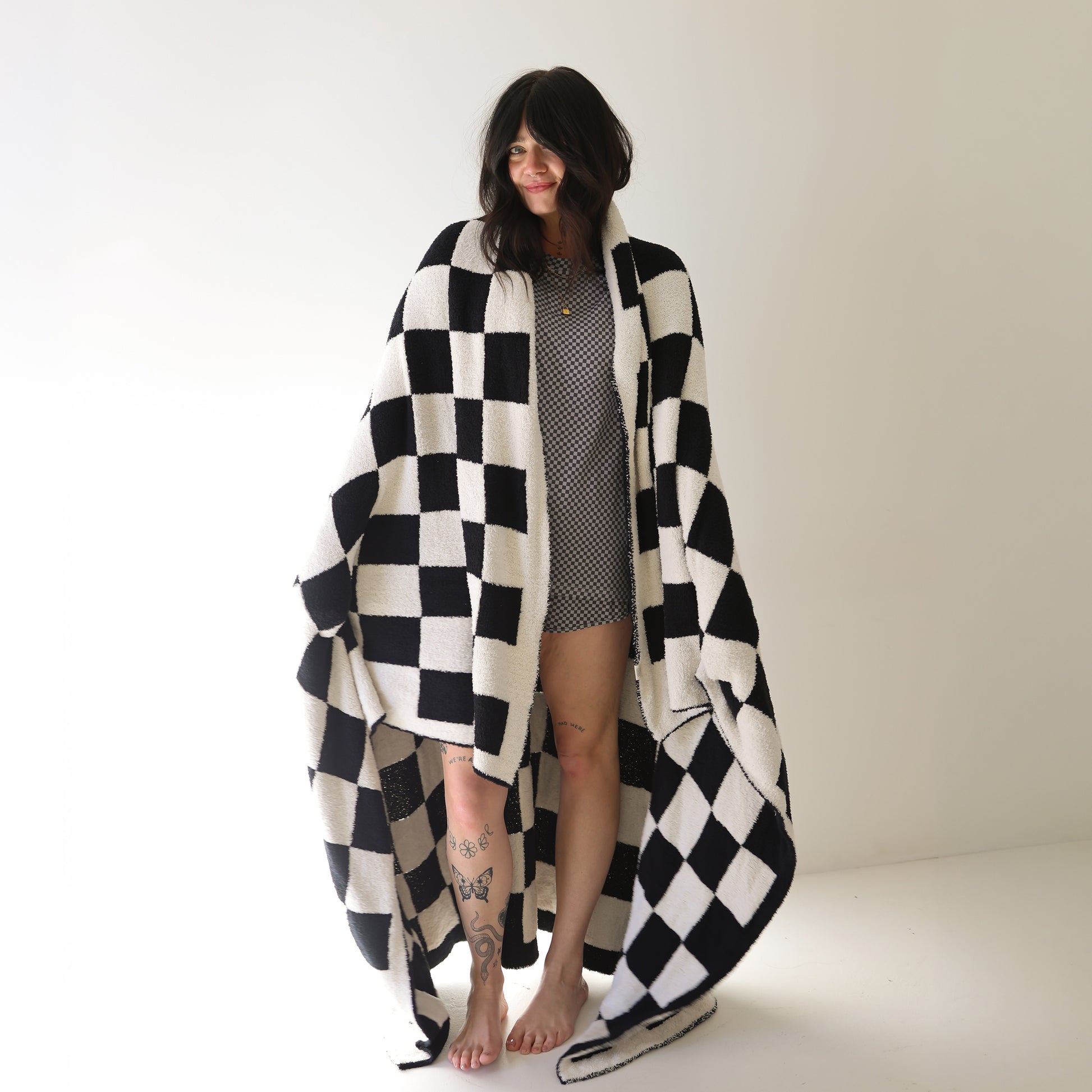 A woman, barefoot in a bright room, is wrapped in the forever french baby Plush Blanket | Black & White Checkerboard. She wears a short patterned dress, has tattoos on her legs, and smiles gently while looking to the side.