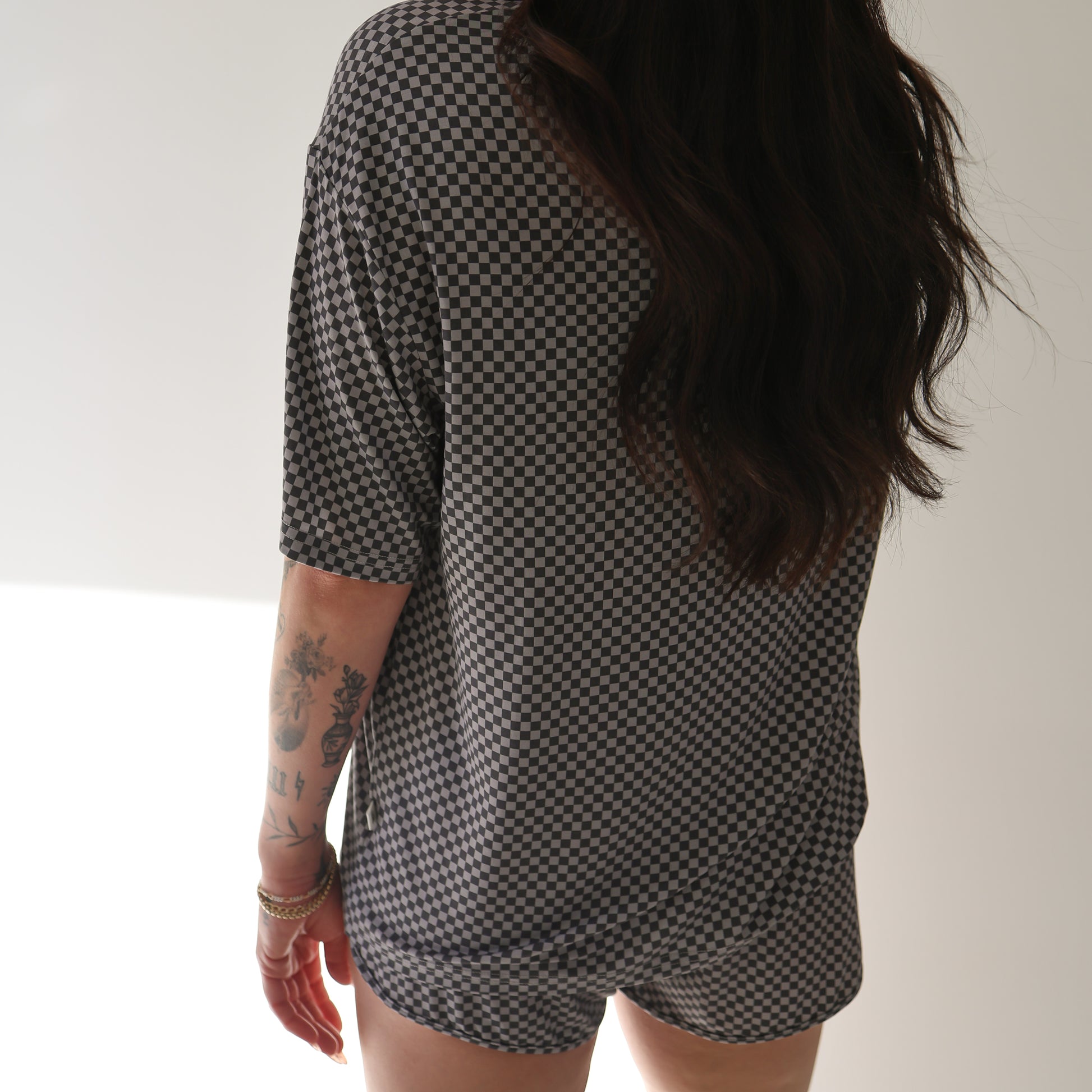 A person with long dark hair, wearing the forever french baby Women's Bamboo Short Set | Street Micro Check, stands facing away from the camera. Hypo-allergenic tattoos are visible on their left arm.