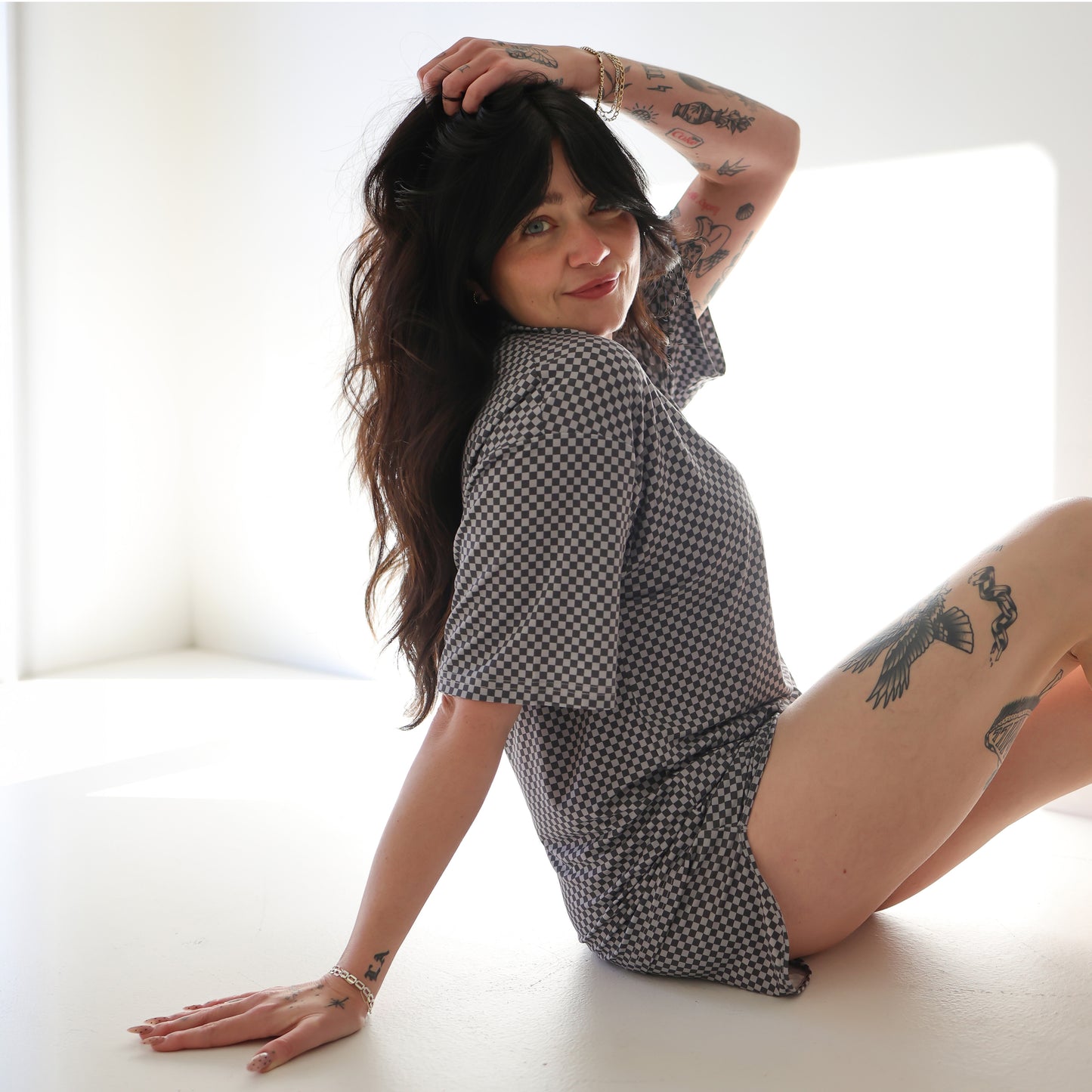 A woman with long dark hair and tattoos smiles at the camera while wearing the forever french baby Women’s Bamboo Short Set | Street Micro Check in a well-lit room.
