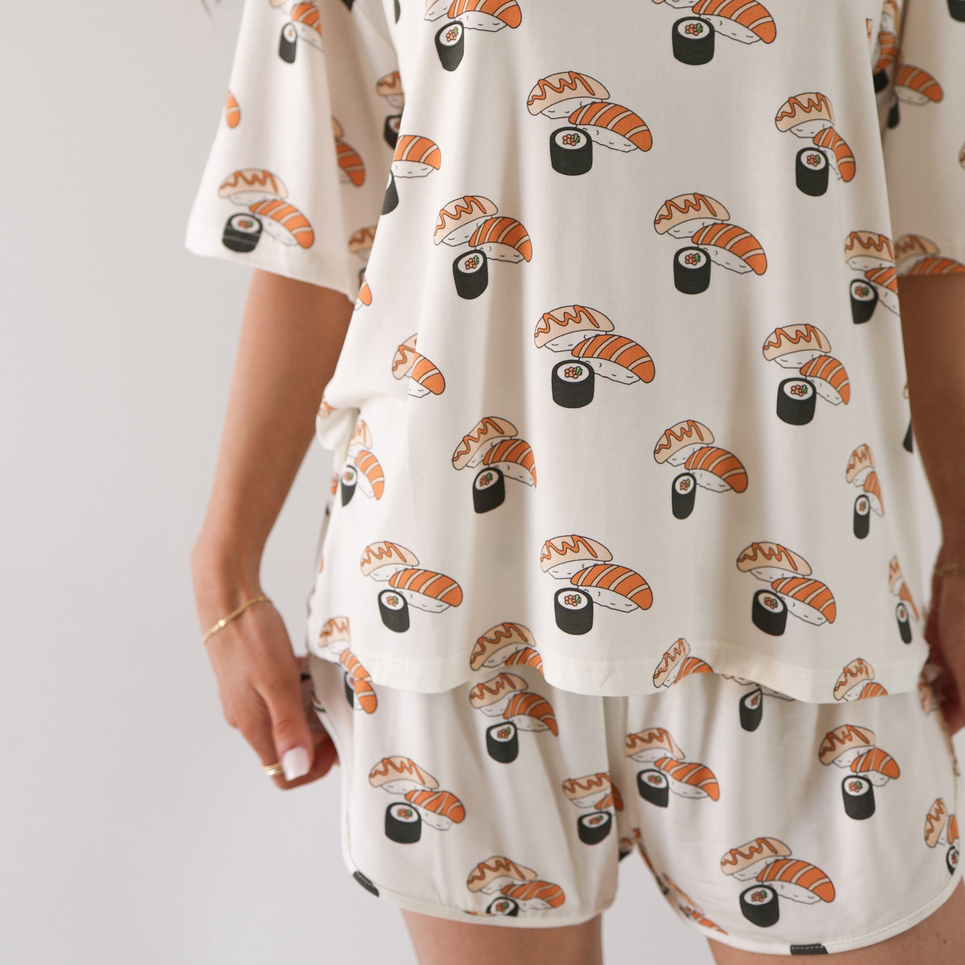 A woman wears the forever french baby Women's Bamboo Short Set | Sushi, a hypo-allergenic loungewear set with matching short sleeves and shorts featuring playful illustrated nigiri and maki, standing against a plain light background.