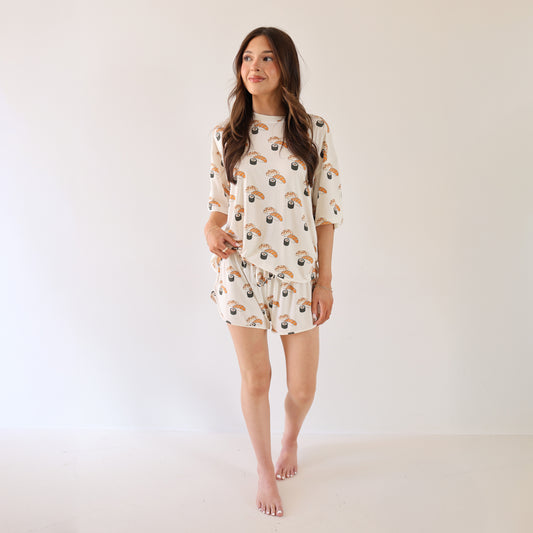 A woman stands barefoot against a white background, wearing the forever french baby Women's Bamboo Short Set in a sushi print. She has long brown hair and smiles gently while looking to the side.