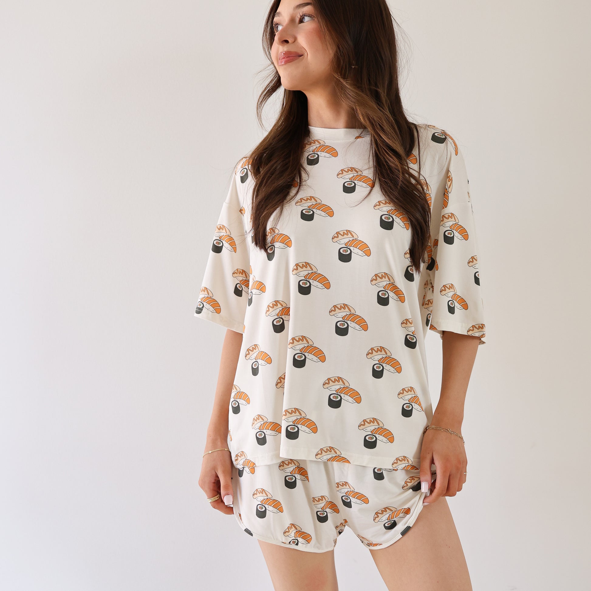 A woman stands against a white background wearing the Women's Bamboo Short Set | Sushi by forever french baby, featuring a matching sushi and soy sauce print with a short-sleeved top and shorts. She looks to the side and smiles slightly.