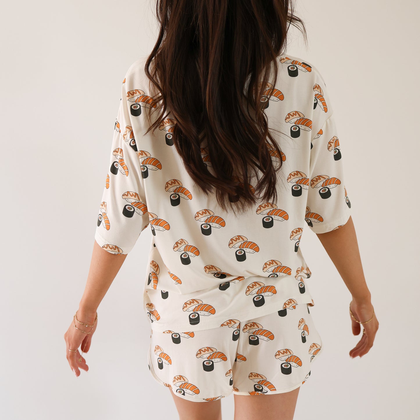 A woman with long brown hair, seen from behind, wears the forever french baby Women's Bamboo Short Set | Sushi, featuring nigiri and maki rolls print, standing against a plain white background.