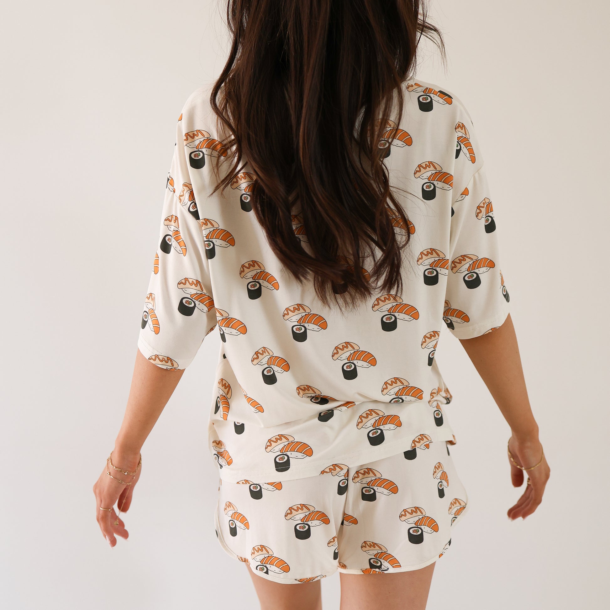 A woman with long brown hair, seen from behind, wears the forever french baby Women's Bamboo Short Set | Sushi, featuring nigiri and maki rolls print, standing against a plain white background.