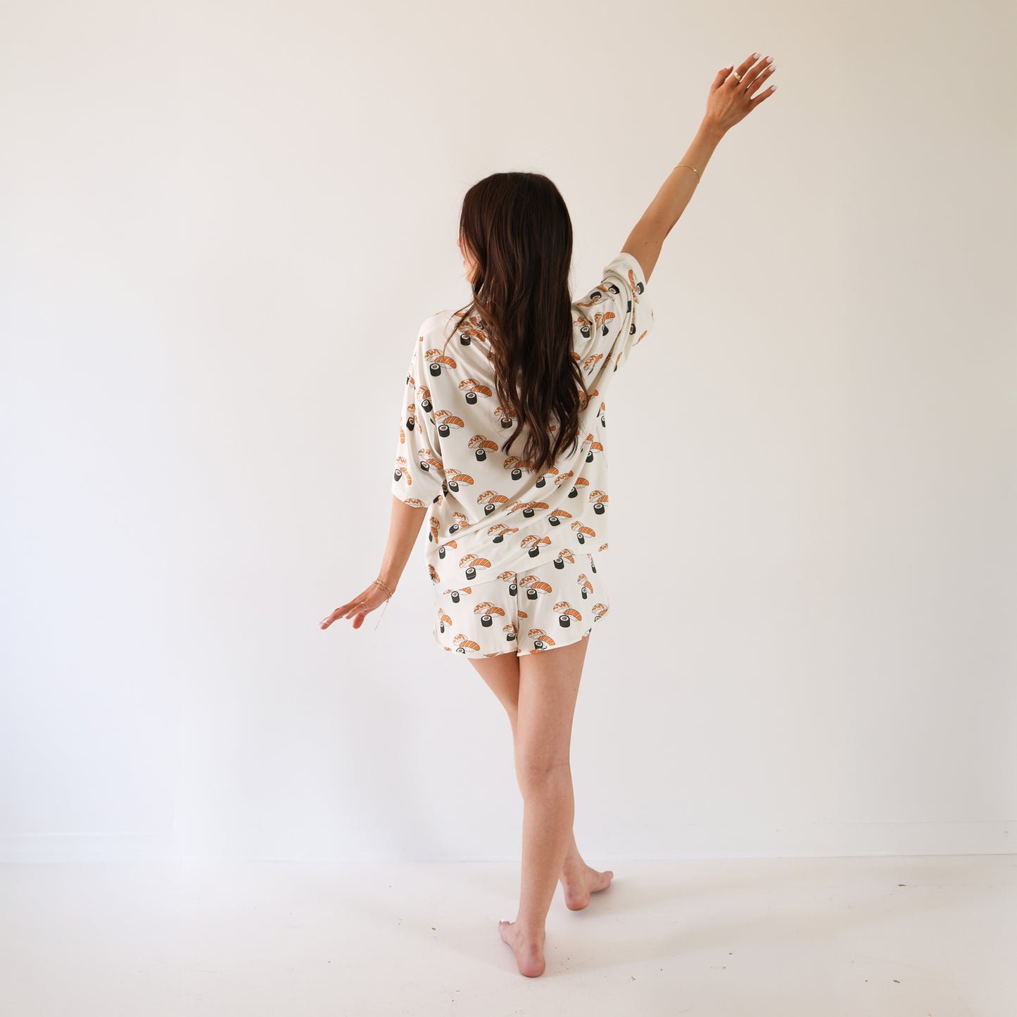A woman with long brown hair stands barefoot in a white room, facing away. She wears the forever french baby Women's Bamboo Short Set | Sushi, featuring a colorful print, and gracefully lifts one arm above her head.