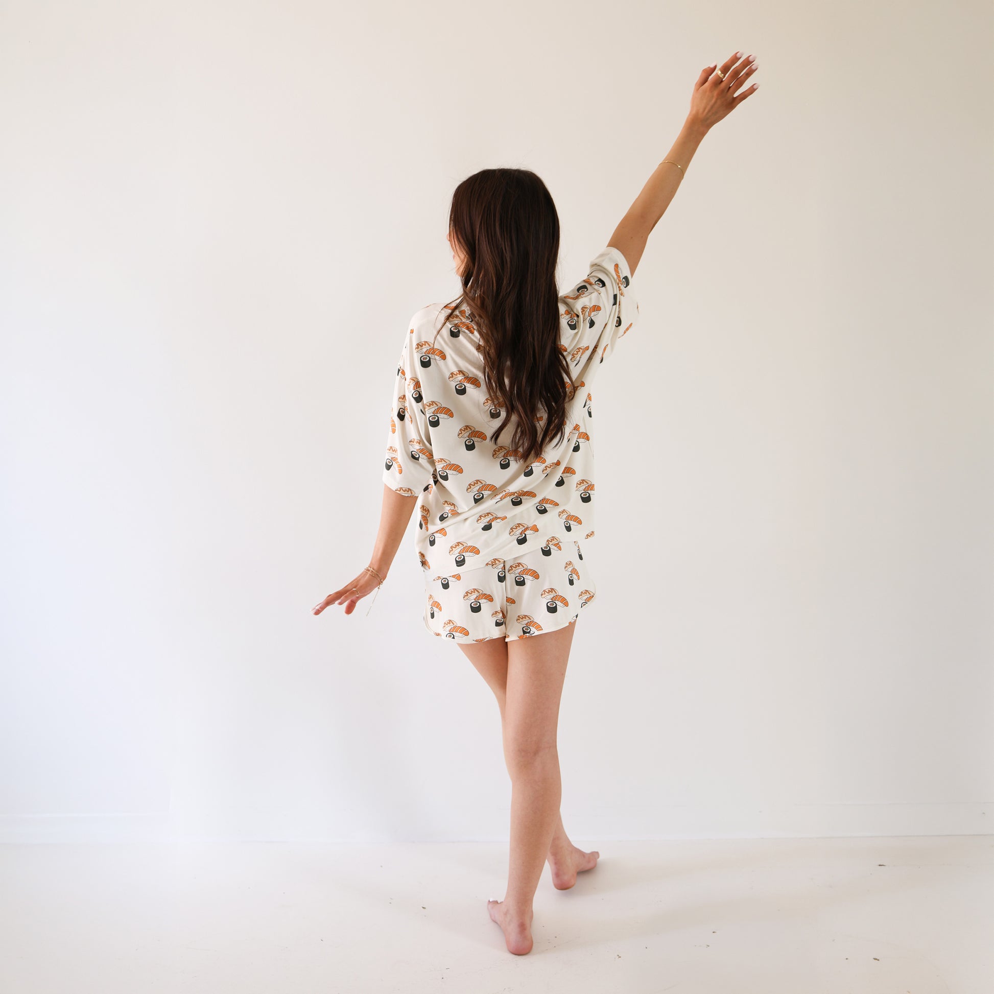 A woman with long brown hair stands barefoot in a white room, facing away. She wears the forever french baby Women's Bamboo Short Set | Sushi, featuring a colorful print, and gracefully lifts one arm above her head.