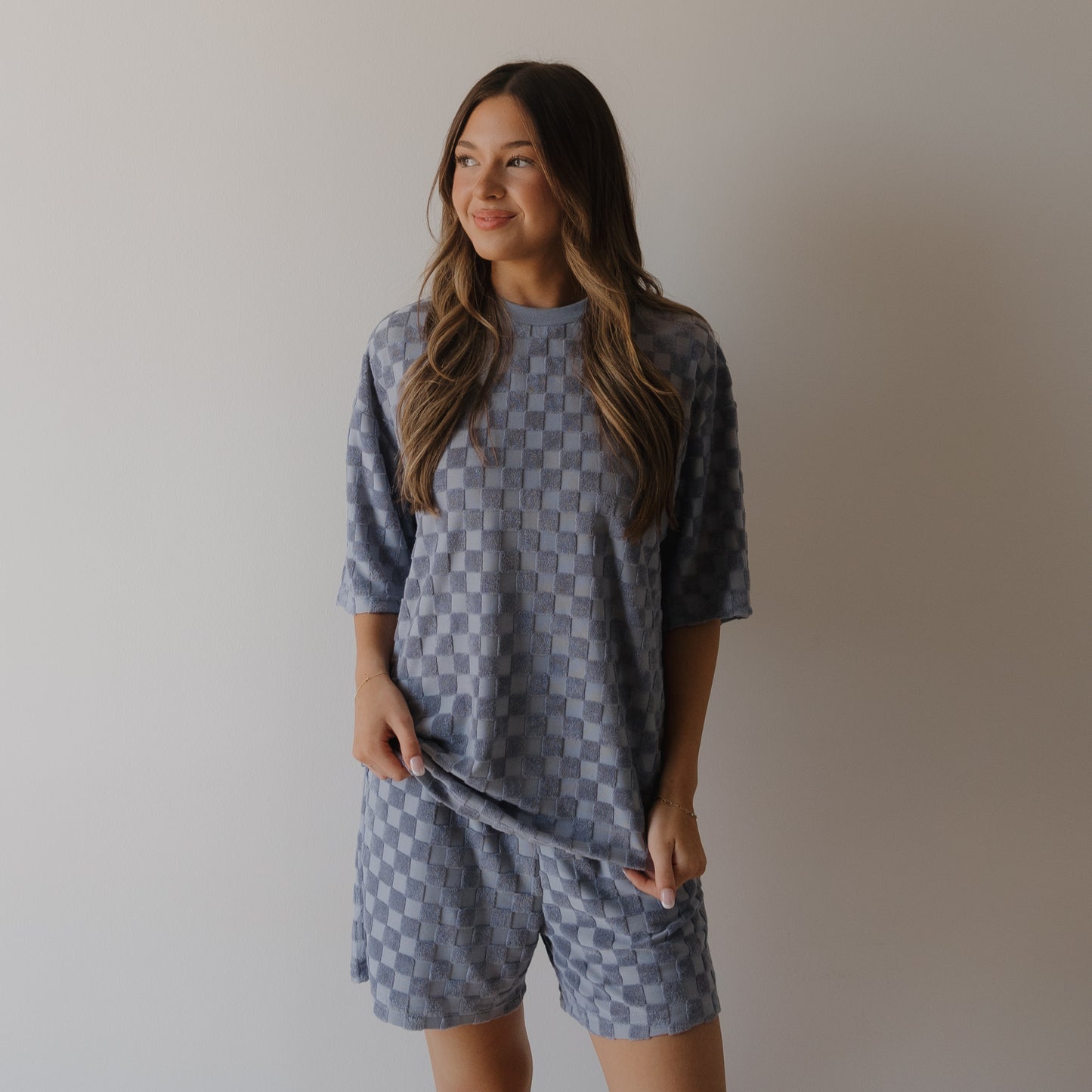 A woman with long, wavy hair stands against a light wall, softly smiling and wearing the forever french baby Women's Terry Short Set in Tradewinds—a loose-fitting blue checkerboard outfit.