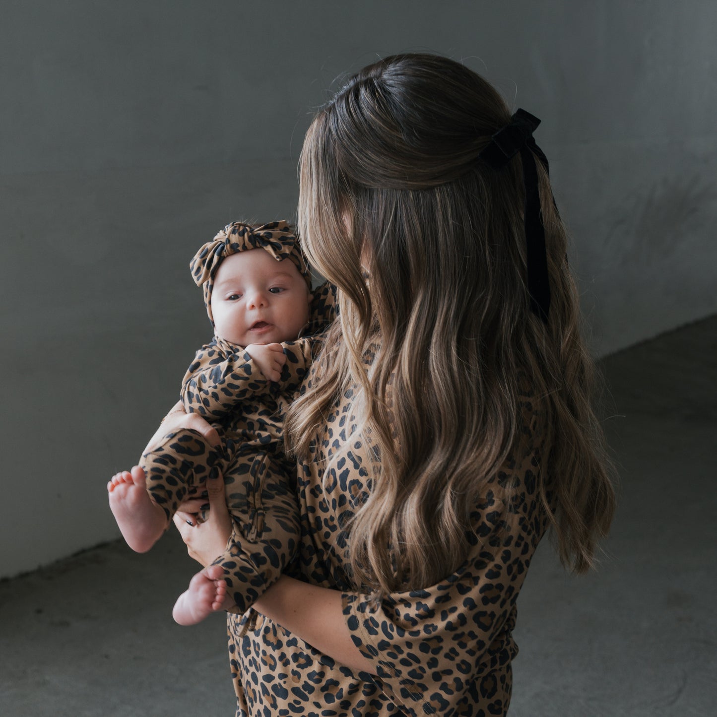 A woman with long wavy hair and a black bow holds her baby as they wear matching forever french baby Bamboo Zip Pajamas in Leopard print. The mom is turned to the side while the baby, in breathable sleepwear, faces the camera.