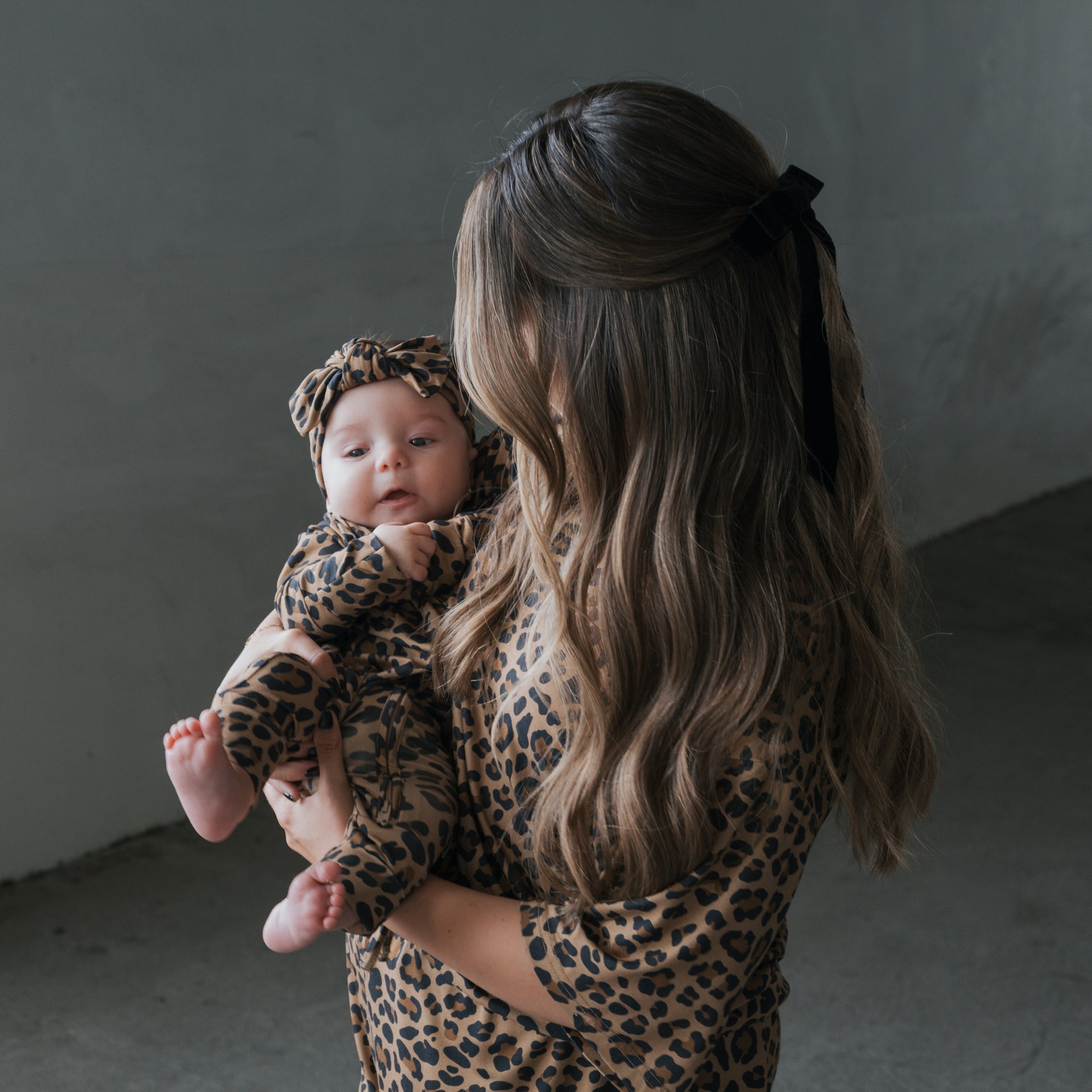 A woman with long wavy hair and a black bow holds her baby as they wear matching forever french baby Bamboo Zip Pajamas in Leopard print. The mom is turned to the side while the baby, in breathable sleepwear, faces the camera.