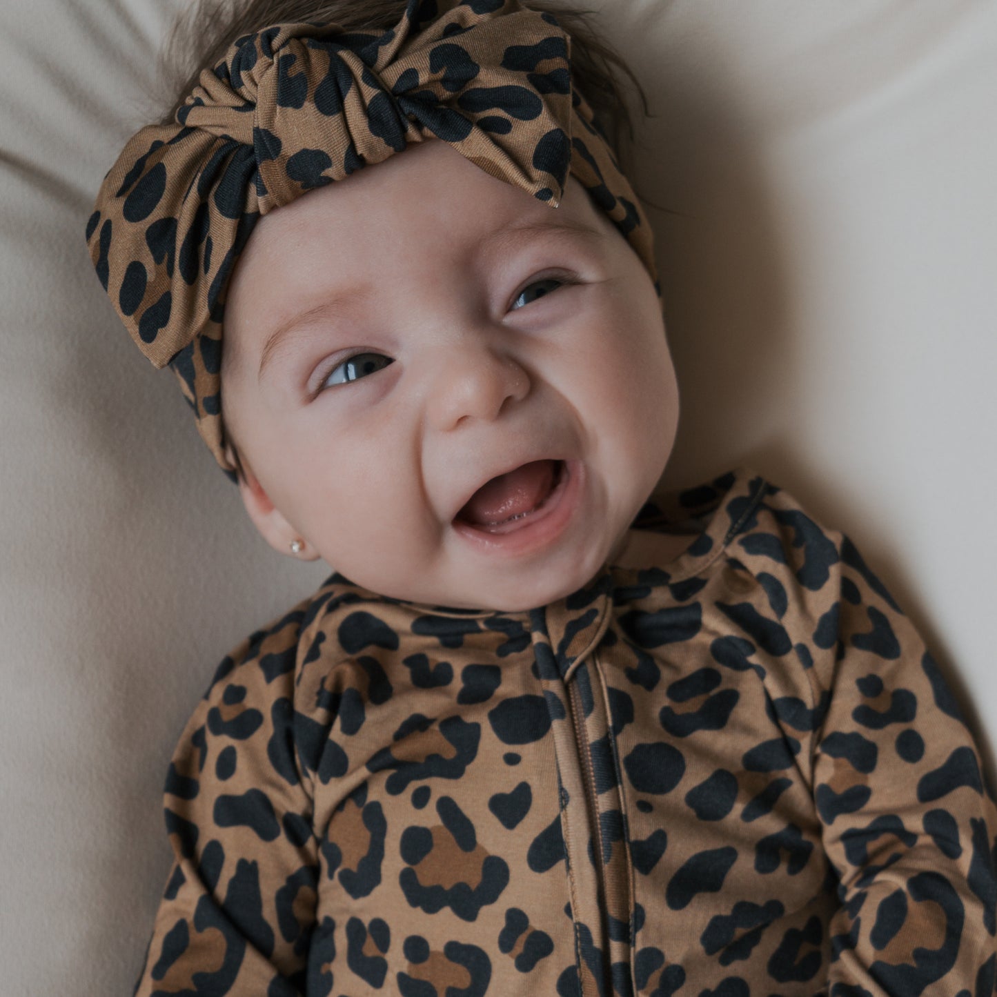 A smiling baby in forever french baby's Bamboo Zip Pajamas | Leopard, featuring a matching headband, lies on a light surface and gazes up with an open mouth and bright eyes.
