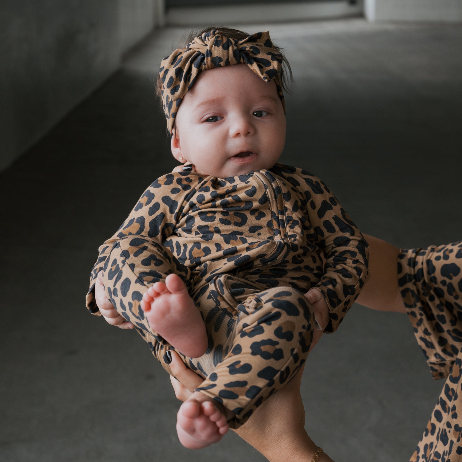 A baby wearing forever french baby Bamboo Zip Pajamas in Leopard print is held by an adult. Barefoot and calm, the baby looks cozy in breathable sleepwear. The dimly lit indoor corridor serves as the background.