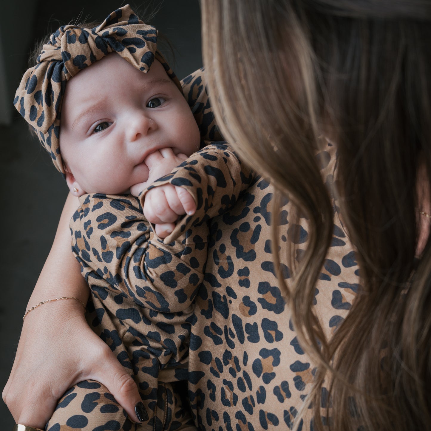A baby in forever french baby's Bamboo Zip Pajamas | Leopard, featuring a matching headband, is held by a woman in coordinated attire. The baby looks up with fingers in their mouth; the woman's face is not visible.