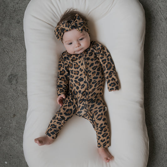 A baby lies calmly on a white cushioned surface, wearing forever french baby's Bamboo Zip Pajamas | Leopard onesie with a matching headband, against a gray textured floor.