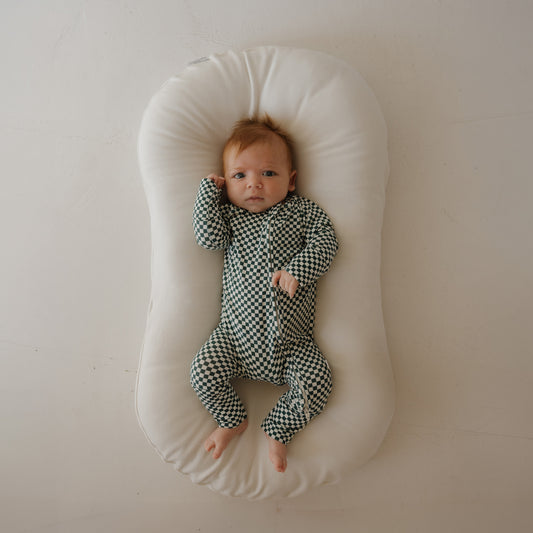 A baby with light brown hair lies on a white, oval cushion, wearing forever french baby's Bamboo Zip Pajamas | Lucky Micro Check—green and white checkered sleepwear—looking up with one arm bent against a plain light background.