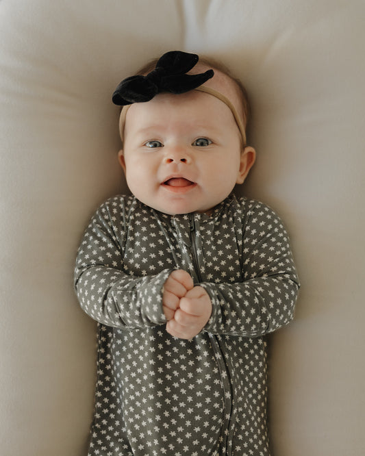 A baby in forever french baby's hypoallergenic Bamboo Zip Pajamas | Green Flowers lies on a light surface, looking up with clasped hands and a slight smile.