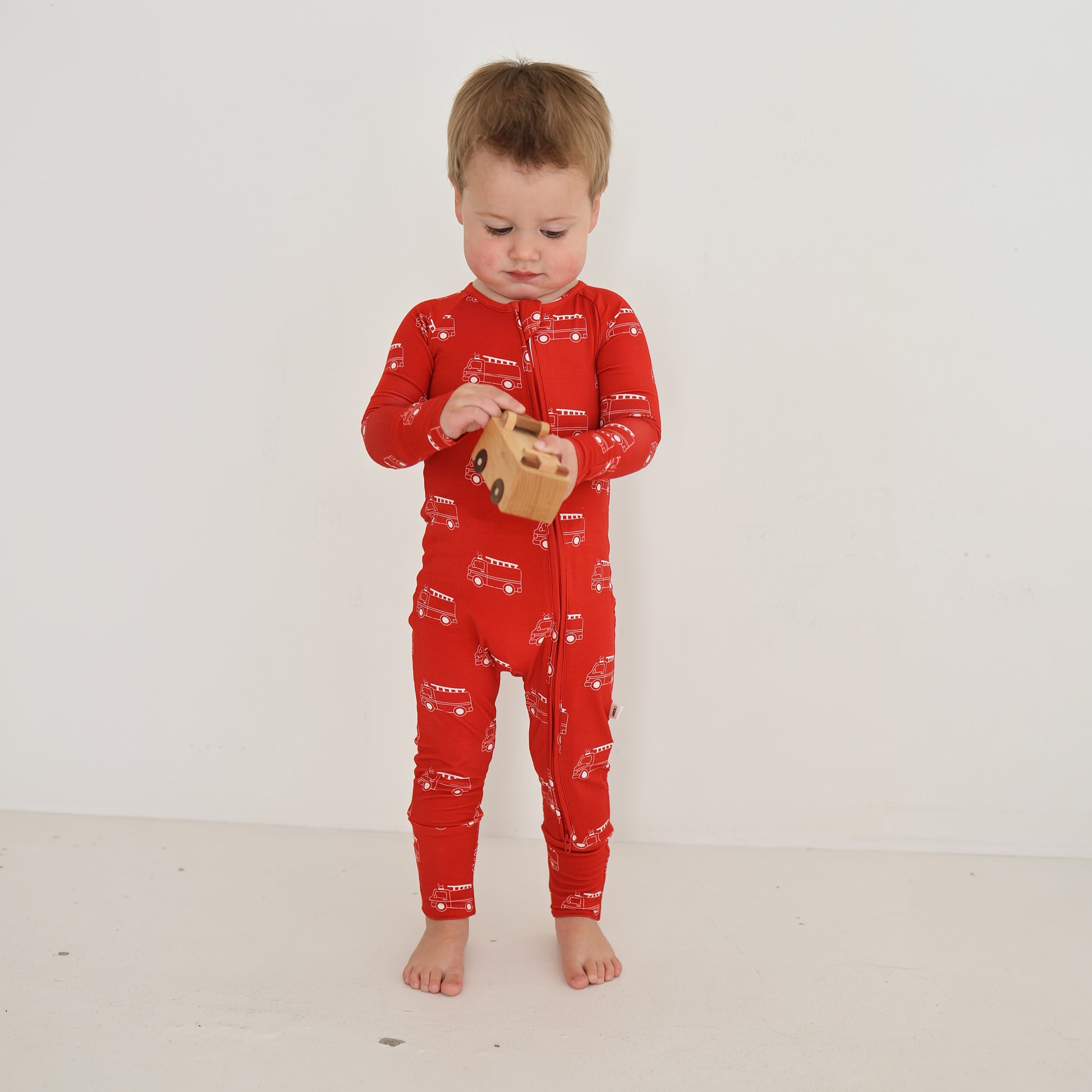A toddler wearing forever french baby Bamboo Zip Pajamas | Fire Truck—a red, breathable onesie—stands barefoot against a white wall, looking down while playing with a small wooden toy.